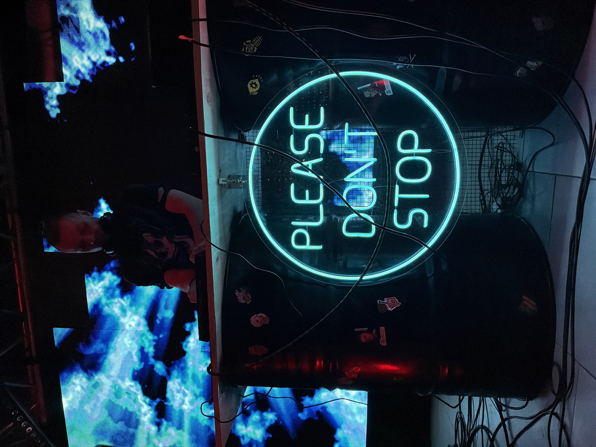 The image shows a DJ booth, likely in a nightclub setting. A person is partially visible behind the equipment, seemingly operating the console. In front of the DJ, a screen displays a blue and white abstract visual effect, resembling flames or flowing water. Above the DJ setup, a circular neon sign displays the words "PLEASE DON'T STOP" in a bright, teal color. The backdrop is dark, with visible speakers, cables, and various stickers attached to the equipment. The specific location within Votkinsk, Russia, cannot be determined from the image alone.