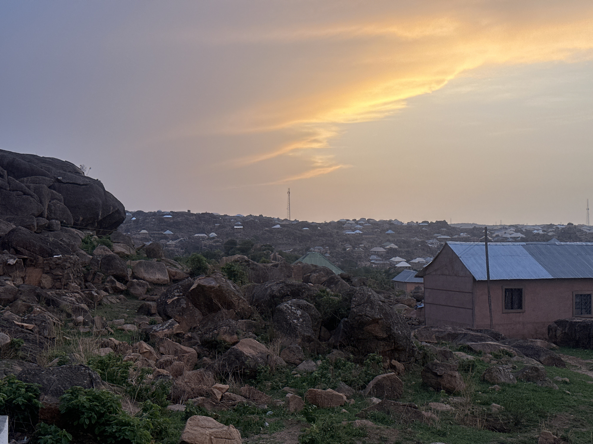 The image depicts a landscape in Jos, Nigeria, characterized by rocky terrain and scattered buildings. A cluster of large, dark rocks dominates the left foreground, interspersed with sparse vegetation. A light brown, single-story building with a corrugated metal roof is visible on the right, with at least two windows and simple architectural details. In the midground, numerous houses with similar roofing materials are spread across the rocky terrain. A tall communication tower stands on the horizon. The sky displays a gradient of colors, suggesting either sunrise or sunset, with yellow and orange hues near the horizon transitioning to a muted gray. The overall lighting appears soft and subdued. No people are visible in the frame.