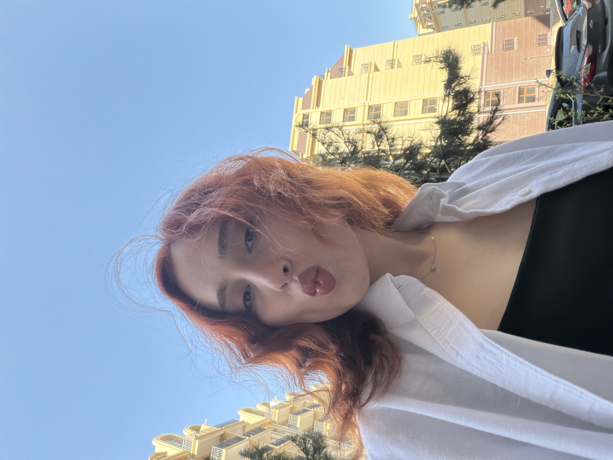 A selfie, captured from a low angle and presented rotated counter-clockwise, features a young person with reddish-brown hair against a clear blue sky in Tbilisi, Georgia. The individual looks towards the camera with a playful "kiss face" expression, lips pursed, and light-colored eyes. They wear a white collared shirt over a black top, with a delicate silver necklace visible on their chest.
The background reveals a vibrant urban setting under bright sunlight. Several multi-story buildings of light yellow or beige hue, featuring windows, balconies, and distinct architectural elements, stand against the clear sky. Dark green trees and shrubs provide greenery amidst the structures. A portion of a dark-colored car is visible on the far right, indicating a street or parking area. The bright, clear sky suggests a sunny morning or afternoon. No text is visible in the image.