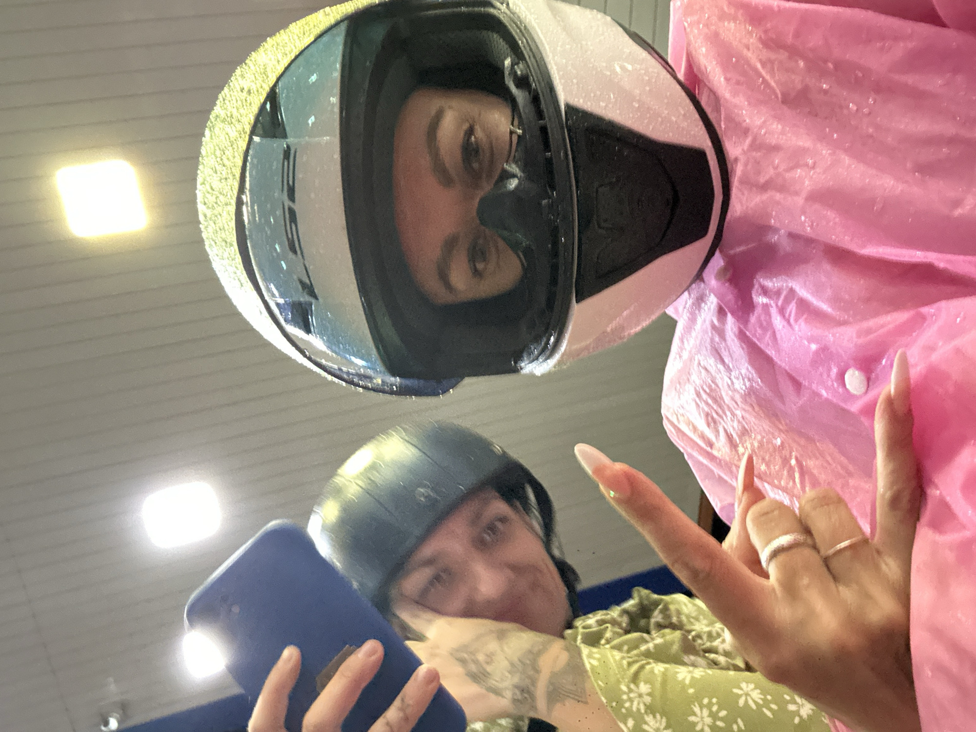 This media file captures two individuals, likely women, from a low-angle perspective looking upwards. The primary subject, positioned at the top of the frame, wears a white and black full-face motorcycle helmet with the brand "LS2" visible on the side. Her eyes are clearly seen through the visor, reflecting a neutral or slightly smiling expression. She is partially covered by a bright pink, wet, and crinkled material, likely a waterproof poncho or raincoat, suggesting recent exposure to rain as water droplets are visible on both the helmet and the pink garment.
The second individual is visible in the lower part of the frame, slightly out of focus. She wears a dark green half-shell helmet and a light green shirt adorned with a white floral pattern, possibly daisies. This person is holding a blue smartphone, from which a bright light or reflection emanates. Her right hand is prominent, making a "rock on" or "I love you" hand gesture, showcasing long, manicured fingernails and two rings on her ring finger.
The background consists of a light-colored ceiling with horizontal paneling and bright rectangular light fixtures, indicating an indoor or sheltered location, possibly a parking garage or covered area. The presence of motorcycle helmets and rain gear is consistent with common modes of transport and weather conditions in a location like Phu Thuy Ward, Vietnam. The overall mood appears casual, despite the implication of wet weather.
