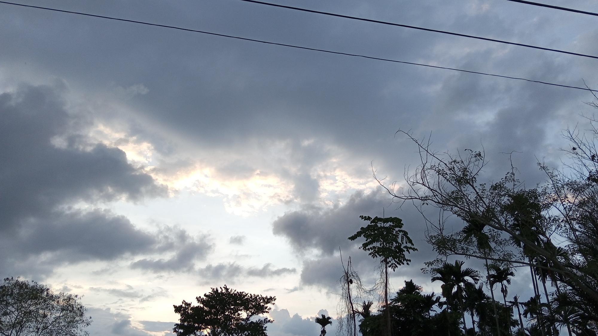 The image depicts a cloudy sky over a landscape in Kilometer Delapan, Indonesia. Dark, grey clouds fill much of the frame, with patches of lighter sky visible in between. In the foreground, there are silhouettes of trees and vegetation. An electrical wire runs across the top of the image.