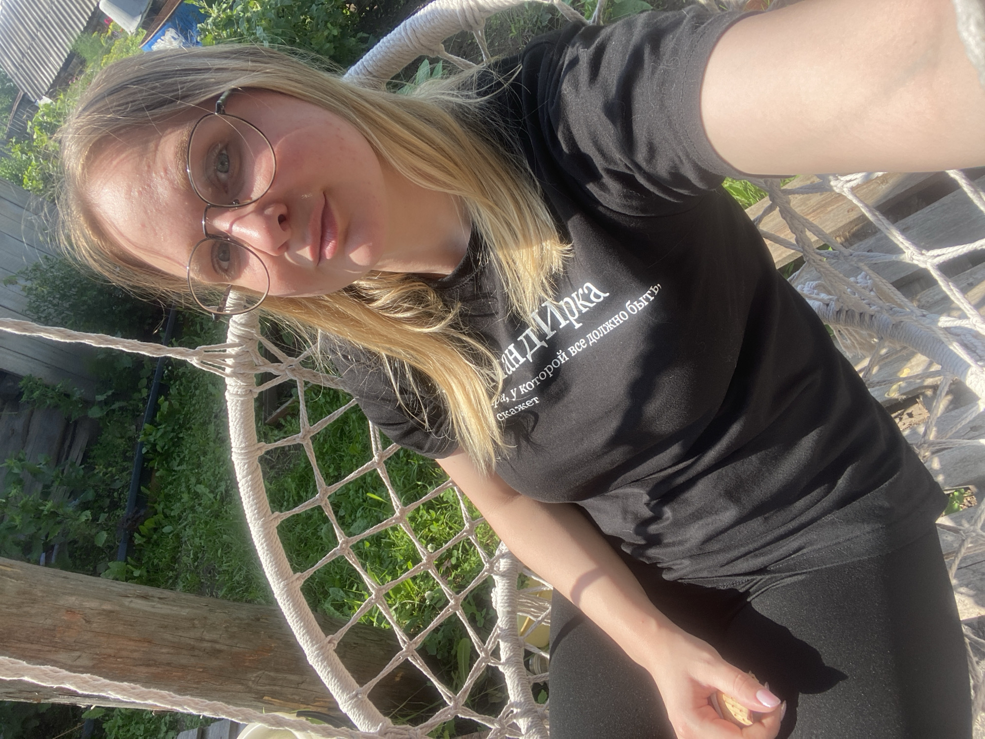A young woman with blonde hair and glasses is sitting in a hanging chair outdoors, holding a cookie in her right hand. She is wearing a black t-shirt with white Cyrillic text that reads, "мандаринка, у которой всё должно быть," which translates to "mandarin, which everything should be." The background shows greenery and a wooden structure, suggesting a rural or backyard setting. The image appears to be a selfie taken on a sunny day. The location of Kauchuk Stantsiya, Russia, is not visually evident from the image itself.