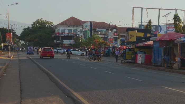 A busy street scene shows a red car driving on an asphalt road. Several motorcycles are also on the road. Shops and buildings line the street, with many people walking along the sidewalks. A large billboard is visible in the background. The sky is bright with sunlight. Trees and foliage are scattered throughout the scene. A lamppost stands tall on the left side of the road.