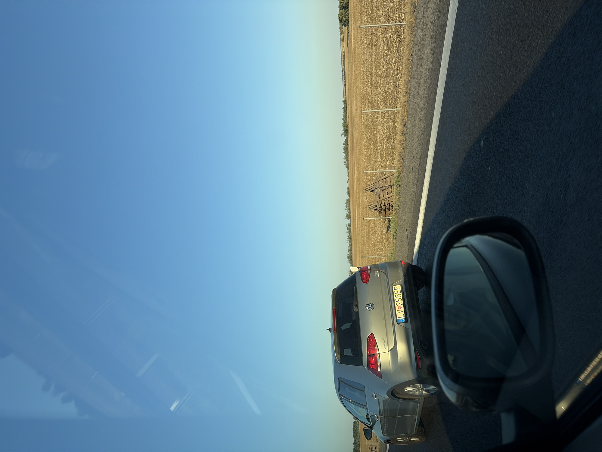 A silver Volkswagen Golf is driving on a highway in Báb, Slovakia. The car's license plate is LV-256EP. The surrounding environment consists of a dry, harvested field with a few trees in the distance. The sky is clear and blue. The image is taken from another vehicle, as evidenced by the reflection in the side mirror.