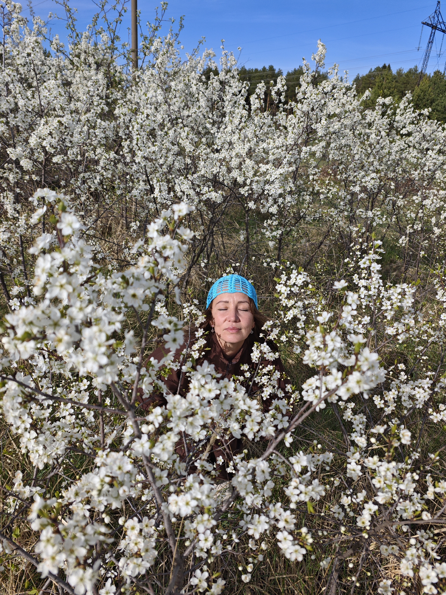 The image depicts a woman with her eyes closed, wearing a blue head covering, positioned within a dense thicket of flowering trees with white blossoms. The scene is likely captured in the city of Chaikovsky, Russia. The background features a clear blue sky, trees, and power lines. The woman appears to be still, potentially meditating or simply enjoying the sensory experience of being surrounded by the blossoms. The flowers are in sharp focus in the foreground and gradually blur into the background. The ground is a mix of grass and brown vegetation.