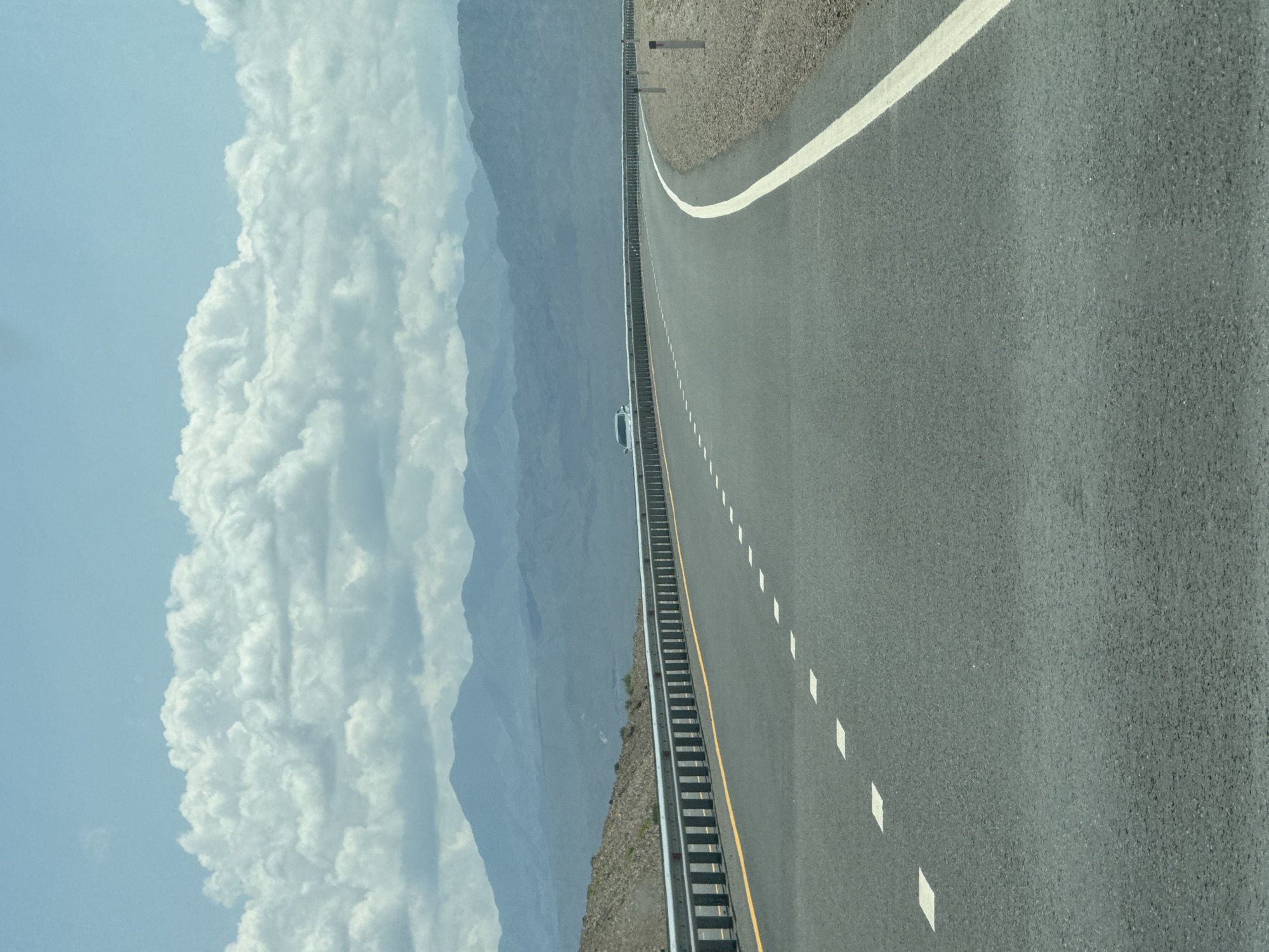This is a medium-angle, eye-level shot of an empty highway curving to the right, suggesting a journey through a desolate, mountainous landscape. The road's asphalt surface is dark gray, with a solid white line on the right edge and a broken white line indicating lanes on the left. A yellow line runs along the shoulder on the far left. A metallic guardrail follows the curve of the road, separating it from a rocky, barren embankment. In the distance, layers of hazy blue mountains rise against a pale blue sky filled with large, fluffy white clouds. A single silver car is visible in the far distance on the road, traveling in the same direction as the viewer. Several small, white posts are intermittently spaced along the right shoulder of the road. The scene appears to be located on a highway leading into or out of the city of Balykchy, Kyrgyzstan, characterized by its vast, open terrain and mountainous backdrop. There are no people visible in the image.