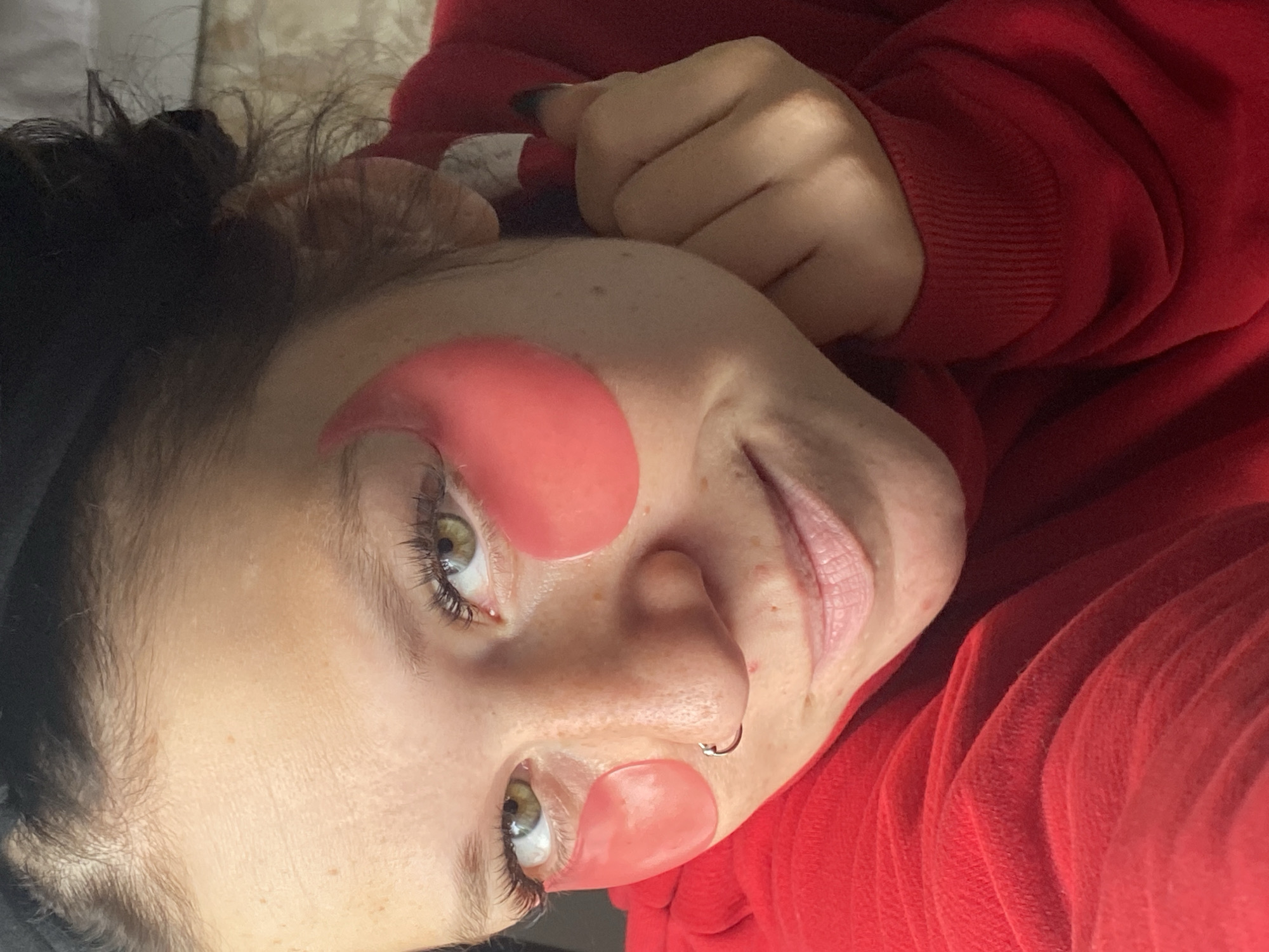 A close-up, over-the-shoulder, selfie-style shot shows a person with fair skin, freckles, and green eyes. The person is wearing pink under-eye masks, has a nose ring, and is wearing a red hooded sweatshirt. Their hair is dark and pulled up. The image appears to have been taken indoors in Chaikovsky, Russia.