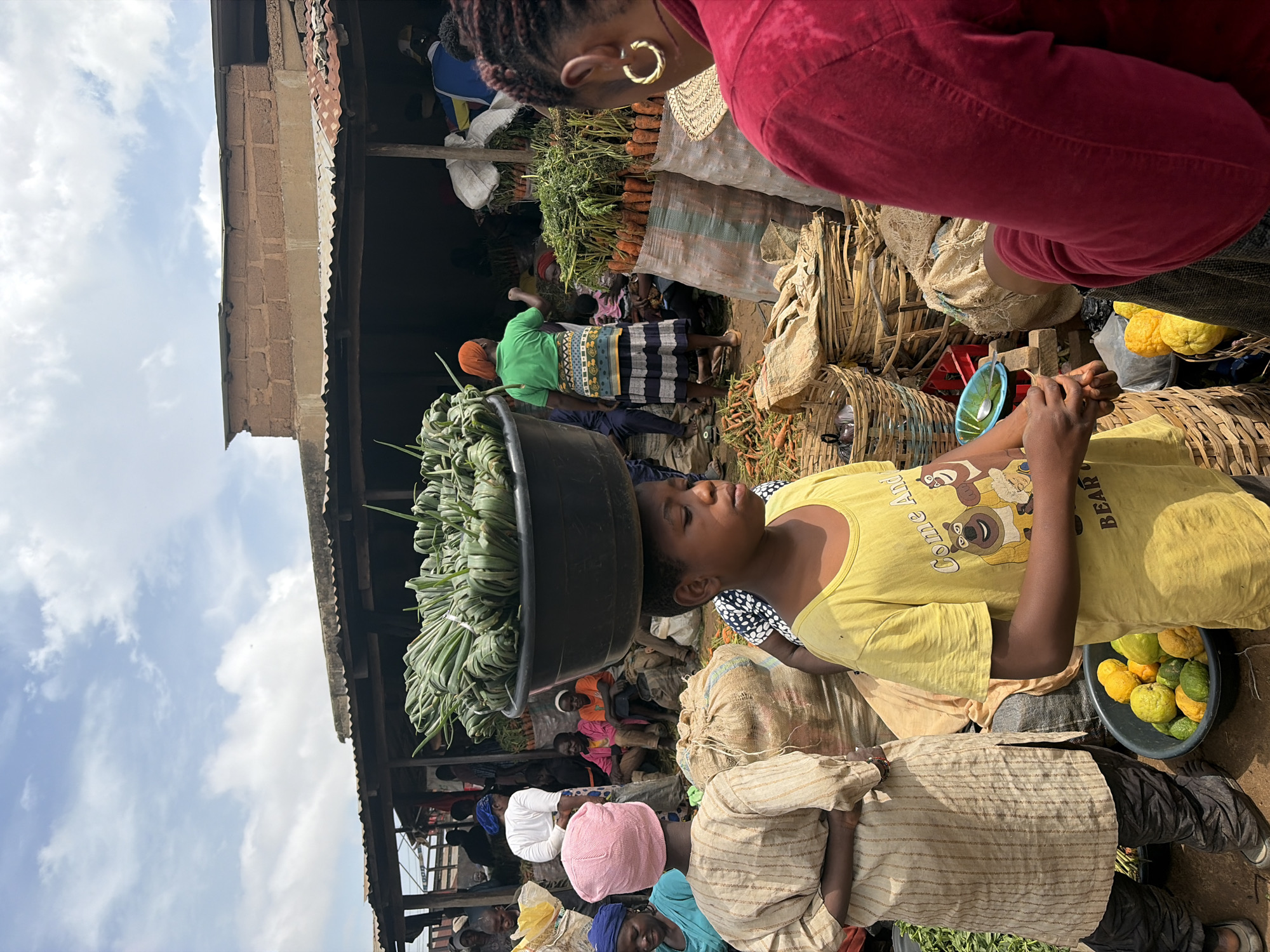 A young boy in a yellow t-shirt carries a large bowl of leafy green vegetables balanced on his head in a bustling outdoor market. He stands in the foreground, facing right, his hands clasped in front of him. Beside him, a person in a striped robe and dark pants faces away from the camera, also appearing to be at the market. In the background, other market-goers are visible, as well as stalls laden with produce, including carrots. The scene takes place in Gura Top, Nigeria, under a partly cloudy sky.