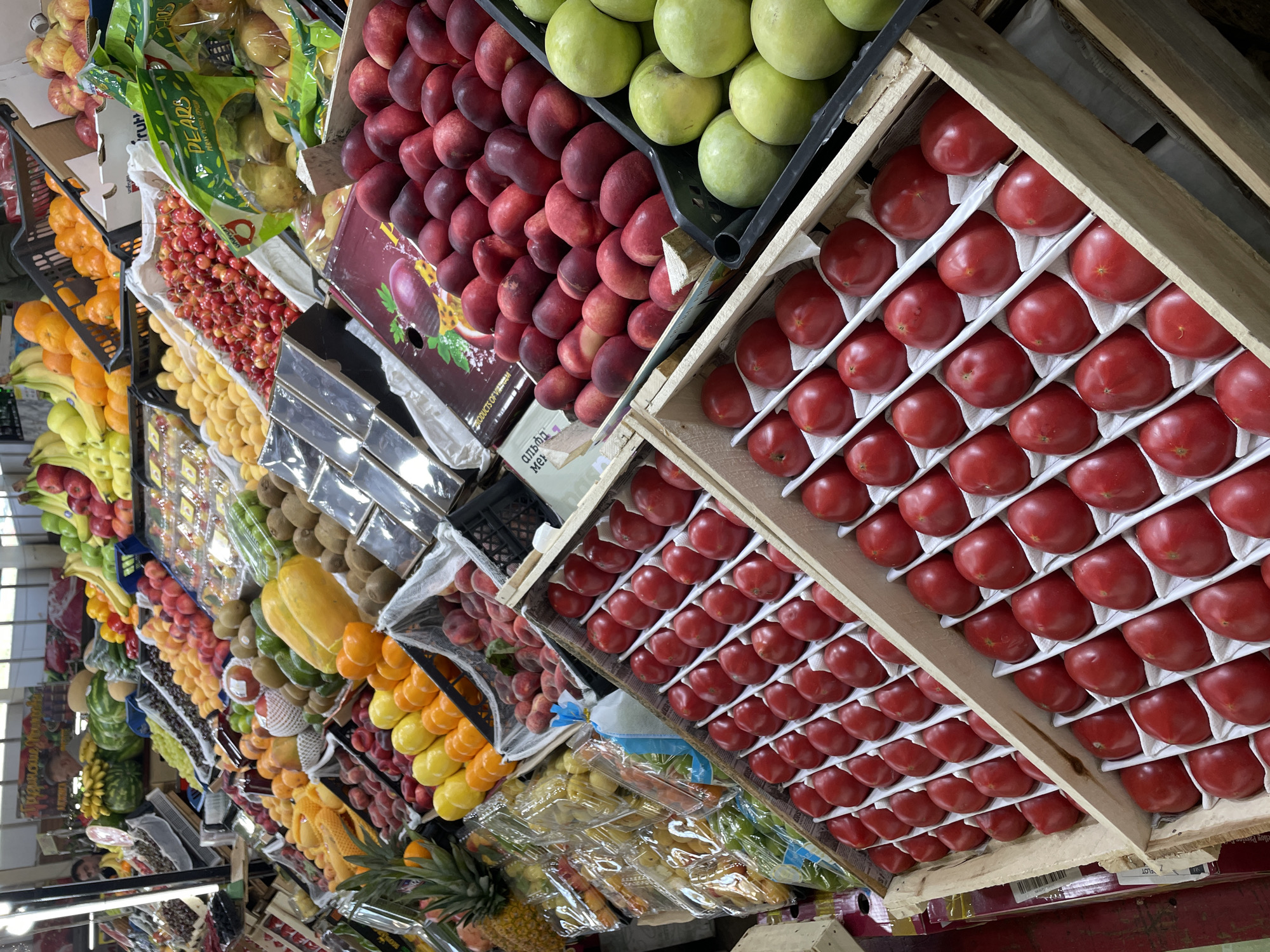 This rotated, close-up image captures a vibrant, well-stocked indoor produce market or fruit stand, showcasing a wide array of fresh fruits and vegetables. The scene is dominated by numerous crates and displays filled with colorful produce, arranged neatly in rows and stacks.
In the foreground and extending to the right, vibrant red tomatoes are meticulously organized in wooden crates, each cushioned by a white paper liner. Above and to their left, large quantities of red peaches or nectarines are piled high, adjacent to bright green apples displayed in black plastic crates. Further left, yellow pears are visible within a green plastic bag labeled "PEARS." Other prominent fruits include piles of red cherries, bright orange oranges, yellow lemons, bananas, kiwis, papayas, and pineapples. Watermelons and what appear to be red apples are also visible towards the left side of the frame. Some produce is packaged in clear plastic containers.
The market appears brightly lit by artificial indoor lighting, highlighting the freshness and variety of the items. A faint reflection of a person is visible in a shiny surface in the lower left, and another person can be seen in the bottom-left corner, suggesting customer or vendor presence.
Visible text includes "PEARS" on a green plastic bag, "АЛЬФА МЕРИ" (Alpha Meri) on a cardboard box, and a partially obscured "PRODUCT OF" on a box featuring artwork of passion fruit. A small label with "LOT" and some numbers is also discernible in the lower right.