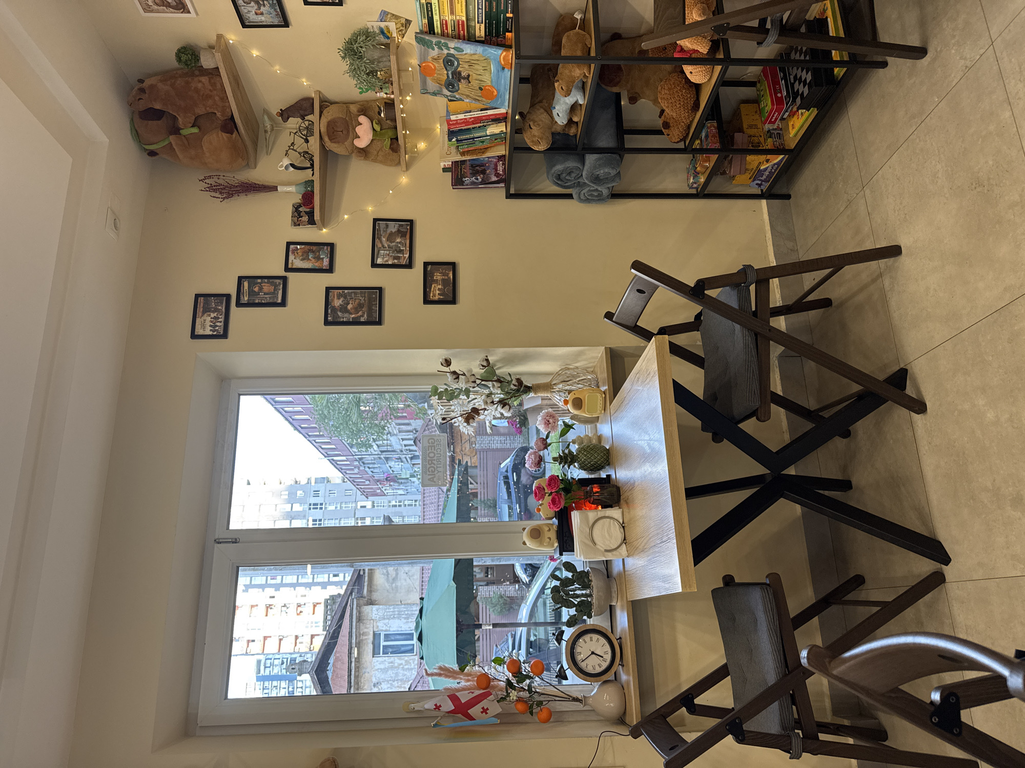 The image captures the interior of a cozy space, potentially a cafe or a room designed for relaxation, situated in an urban environment. A large window provides a view of a bustling street, complete with buildings, parked cars, and glimpses of vegetation, suggesting a central location within a city.
The interior is decorated with an eclectic mix of items. On the wall, shelves display stuffed animals, books, and other decorative objects. Additionally, a string of lights adds a warm ambiance to the room. Below the shelving, several framed pictures are arranged on the wall.
A light-colored wooden table is positioned in front of the window, adorned with an assortment of decorative items including flowers, a clock, and additional ornaments. Several dark wooden chairs are arranged around the table, indicating a seating area designed for conversation or quiet contemplation. The scene suggests a blend of comfort and visual interest, typical of spaces designed to foster a welcoming atmosphere.