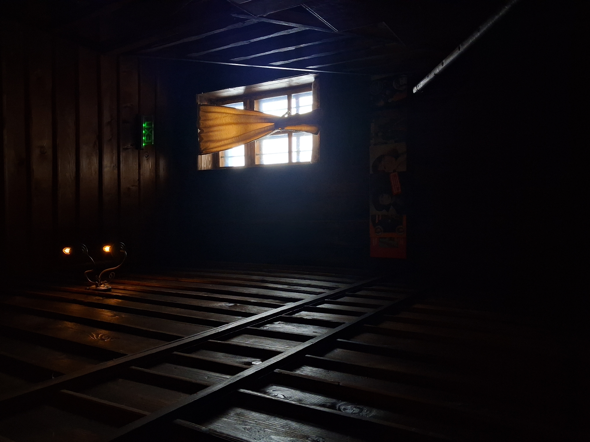 The image depicts a dimly lit interior space with a rustic, possibly historical, atmosphere. The room appears to be constructed of wood paneling and beams. A window is visible, admitting a significant amount of light, with a yellow curtain tied in the middle. There's an 'EXIT' sign to the left of the window. A track-like structure runs across the wooden floor. There are two small lamps near the wall to the left of the window, adding some artificial illumination. The objects hanging on the right wall are covered in darkness. The location is Katunishte, Bulgaria.