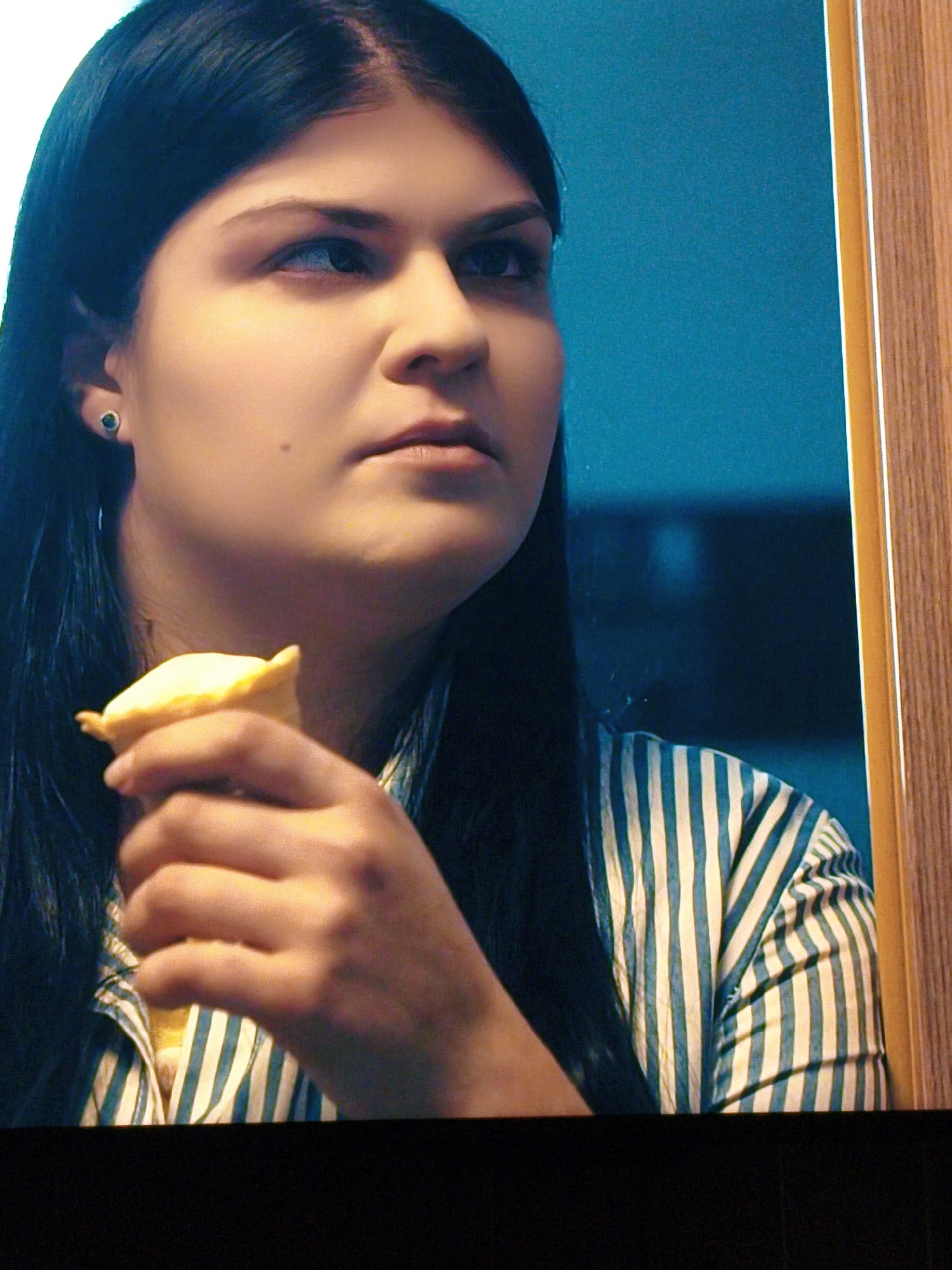The image shows a close-up of a young woman with dark hair, wearing a blue and white striped shirt. She is holding a yellow, cone-shaped pastry to her mouth. Her left earring is visible, a small dark stud. The background is a blurred teal wall, with a hint of a wooden frame on the right side of the image. The scene appears to be indoors, possibly a kitchen or dining area. There is no specific information or visual cues in the image to suggest a location in Vladivostok, Russia.