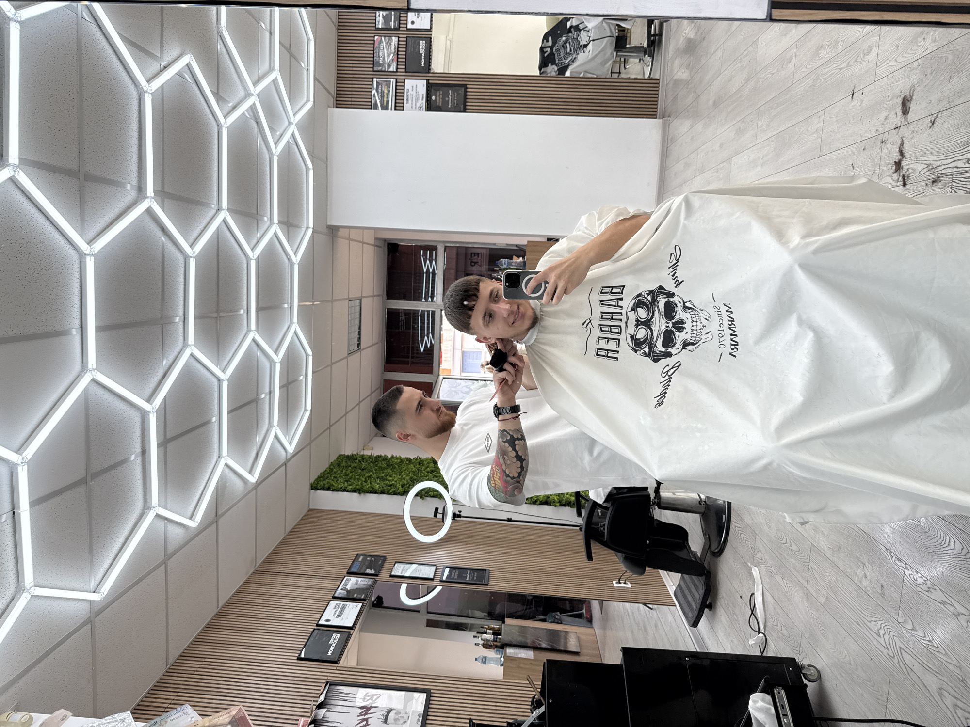 The image captures a scene inside a barbershop. A man with a short haircut is holding up a smartphone to take a selfie while wearing a white barber cape with a skull graphic on the front. Standing behind him is a barber with a beard and a tattooed arm, holding clippers as if in the process of giving the man a haircut.
The environment includes a grey wood-patterned floor, a wall with hexagonal neon lights, and wood paneling. A ring light can be seen on a stand. Certificates or licenses are framed and hung on a wall. The reflection in the mirror shows part of the interior of the barbershop. Based on the reflection of the entrance/exit in the mirror, it appears to be in the city of Sofia, Bulgaria, due to Cyrillic script visible.
