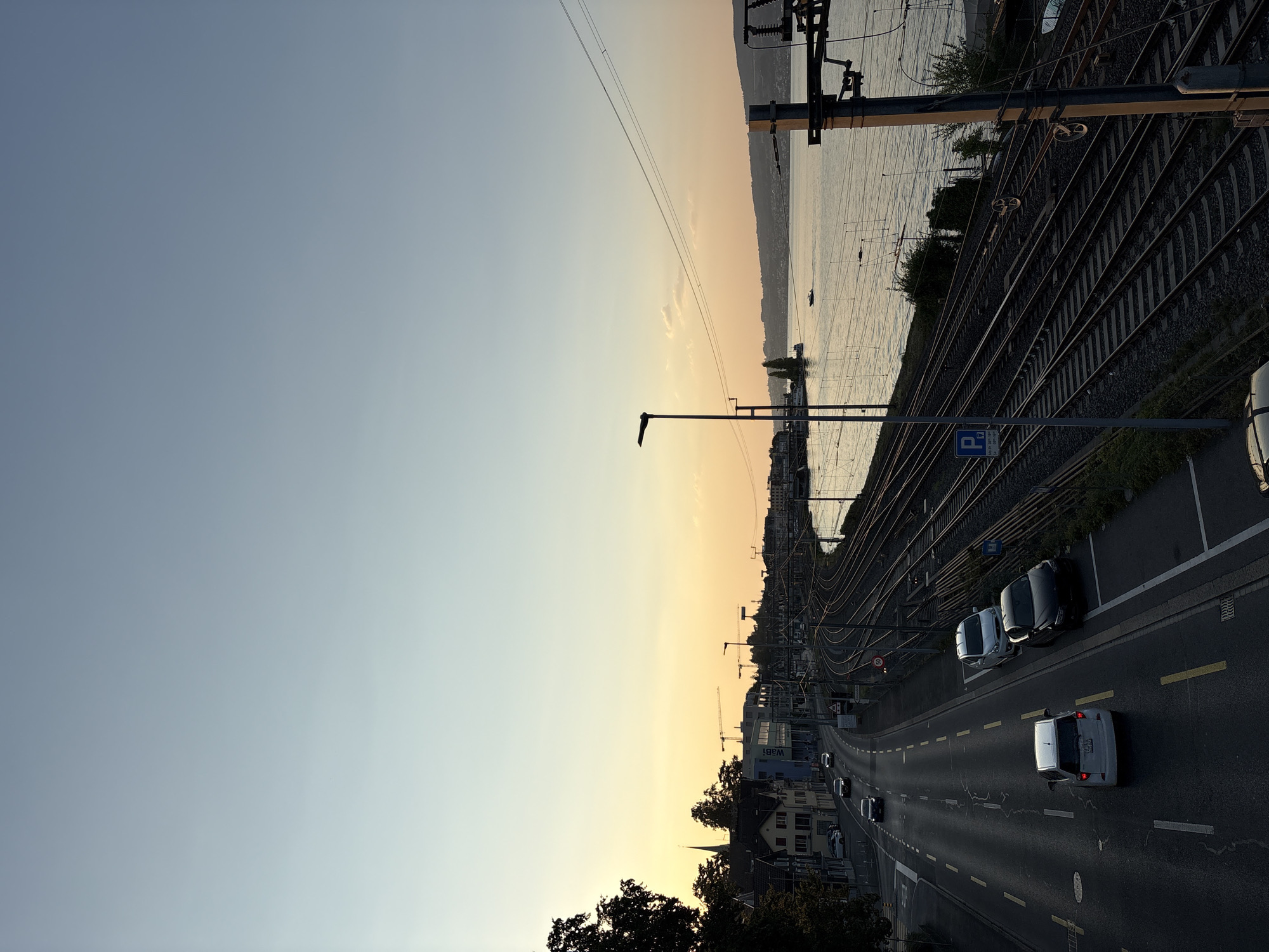The image depicts a sunset view of the city of Wädenswil, Switzerland, featuring a prominent lake, railway tracks, and a road. In the foreground, a road with lane markings is visible, with several cars driving and parked. A white hatchback is in the left lane, followed by a dark-colored sedan in the right lane. Further down the road, a white SUV and a dark sedan are parked on the right side. To the right of the road, an extensive network of railway tracks runs parallel to the water. A blue parking sign is visible near the tracks.
The lake, likely Lake Zurich, occupies the middle and right portions of the image, reflecting the warm hues of the sunset. A small boat is visible on the water. The horizon is marked by a distant shoreline with hills. On the left side of the image, buildings characteristic of a town or city line the road, including houses and a modern building with the name "wabi" visible. A church spire can be seen among the buildings. Trees are present on the far left, casting shadows. The sky is a gradient of light blue and soft orange, indicative of dusk. Electrical poles and wires are present, particularly near the railway tracks. The scene is captured from an elevated perspective.