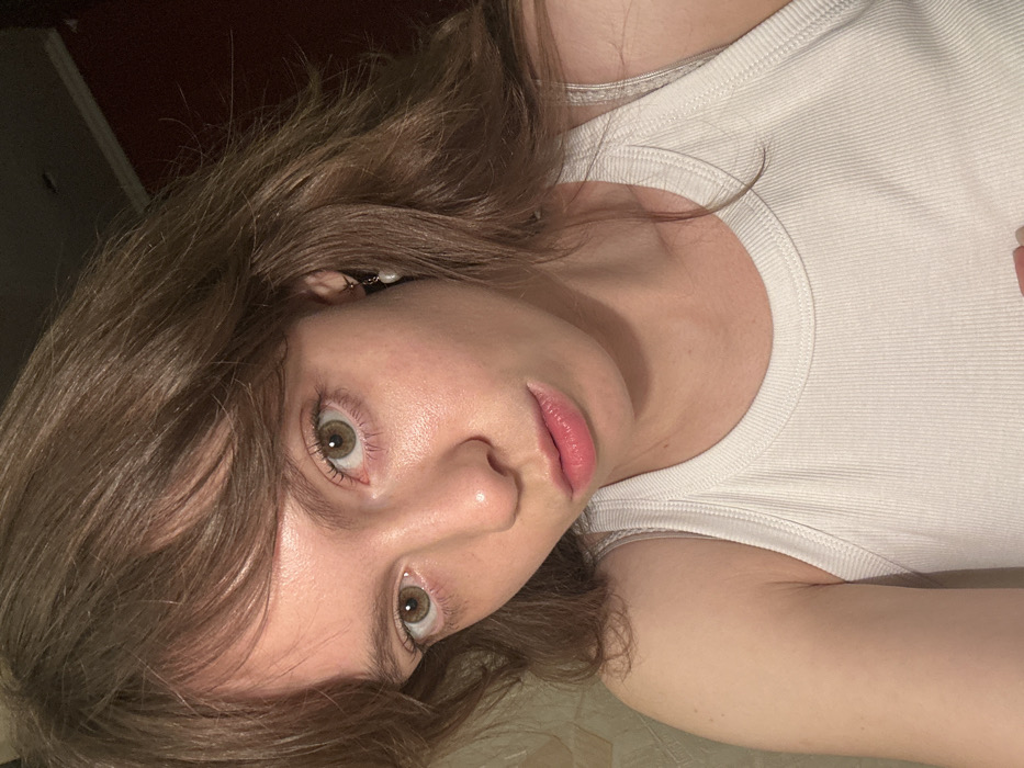 The image displays a close-up, head-and-shoulders portrait of a young adult female with fair skin, light hazel-green eyes, and long, wavy brown hair with a fringe. She is looking directly at the viewer with a neutral expression. Her lips are naturally pink and slightly parted. She is wearing a white ribbed tank top. A small, gold-toned hoop earring with a single white pearl-like drop is visible on her right ear. The background is indistinct, showing a light-colored, possibly textured wall section on the left and a darker, solid reddish-brown or maroon surface to the right and behind her. The lighting appears to be direct, possibly from a flash. The specific location within the city of Moscow, Russia, cannot be determined from the visual information present in the image.