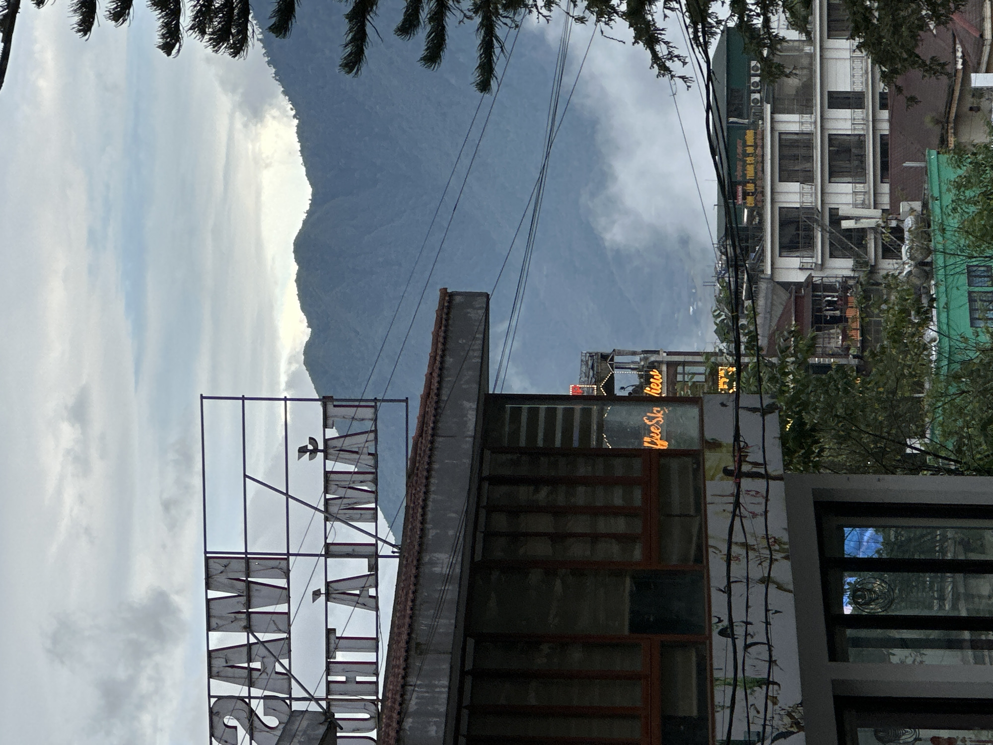The image depicts a scene in Sa Pa Ward, Vietnam, featuring a mountain backdrop partially obscured by clouds. Foreground elements include a large sign that reads "SAPALY HOTEL," indicating the location. Adjacent to the hotel sign is a building with windows, and a neon sign above that reads "Coffee Sky View." In the background, a multi-story building with a green roof can be seen. Power lines are strung across the view. A few trees appear at the top and right of the frame.