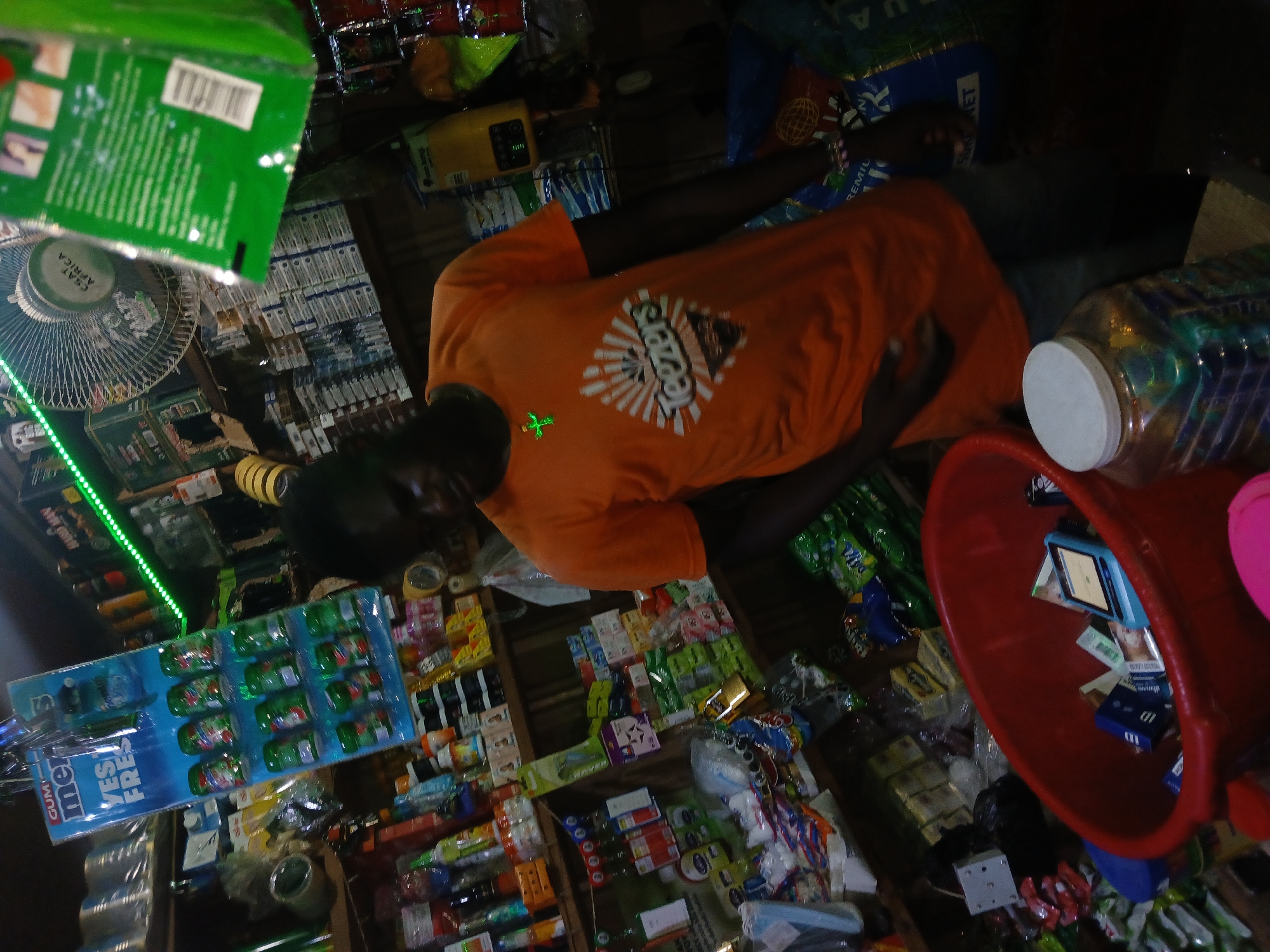 The image depicts an indoor market stall, likely in Jalingo, Nigeria, filled with various goods. A young Black male wearing an orange t-shirt with a graphic print and blue jeans stands in the center. He is looking downwards, with his right hand partially obscuring his face. On his t-shirt, the word "Teezers" is visible in large white lettering. He is wearing a green cross-shaped pendant.
The stall is densely packed with products displayed on shelves and hanging from the ceiling. Prominently featured is a display of "GUM MERRIES" chewing gum packs. Other items visible include cleaning supplies, packaged food items, and various plastic containers. A red plastic basin is in the foreground, containing what appears to be cigarette packs and a blue electronic device. To the right of the basin is a large, clear plastic jar filled with colorful items, possibly candies or small toys. A ceiling fan is visible in the upper right portion of the image, and a green LED light strip runs along the top of the stall. The overall lighting is dim, with some artificial light sources contributing to the scene. The environment suggests a busy retail setting within Jalingo.