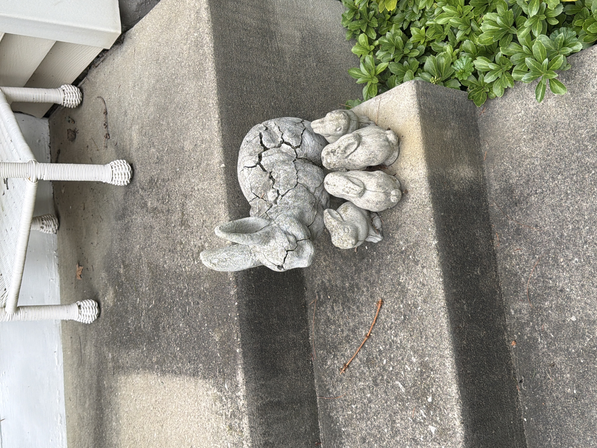 A concrete garden statue of a mother rabbit with four smaller rabbits sits on a gray concrete step outside a building. The mother rabbit is cracked and weathered. The smaller rabbits are clustered around her. Lush green foliage is visible to the right of the step. A white wicker chair is partially visible at the top of the steps. The location is North Beaver Township, United States of America.