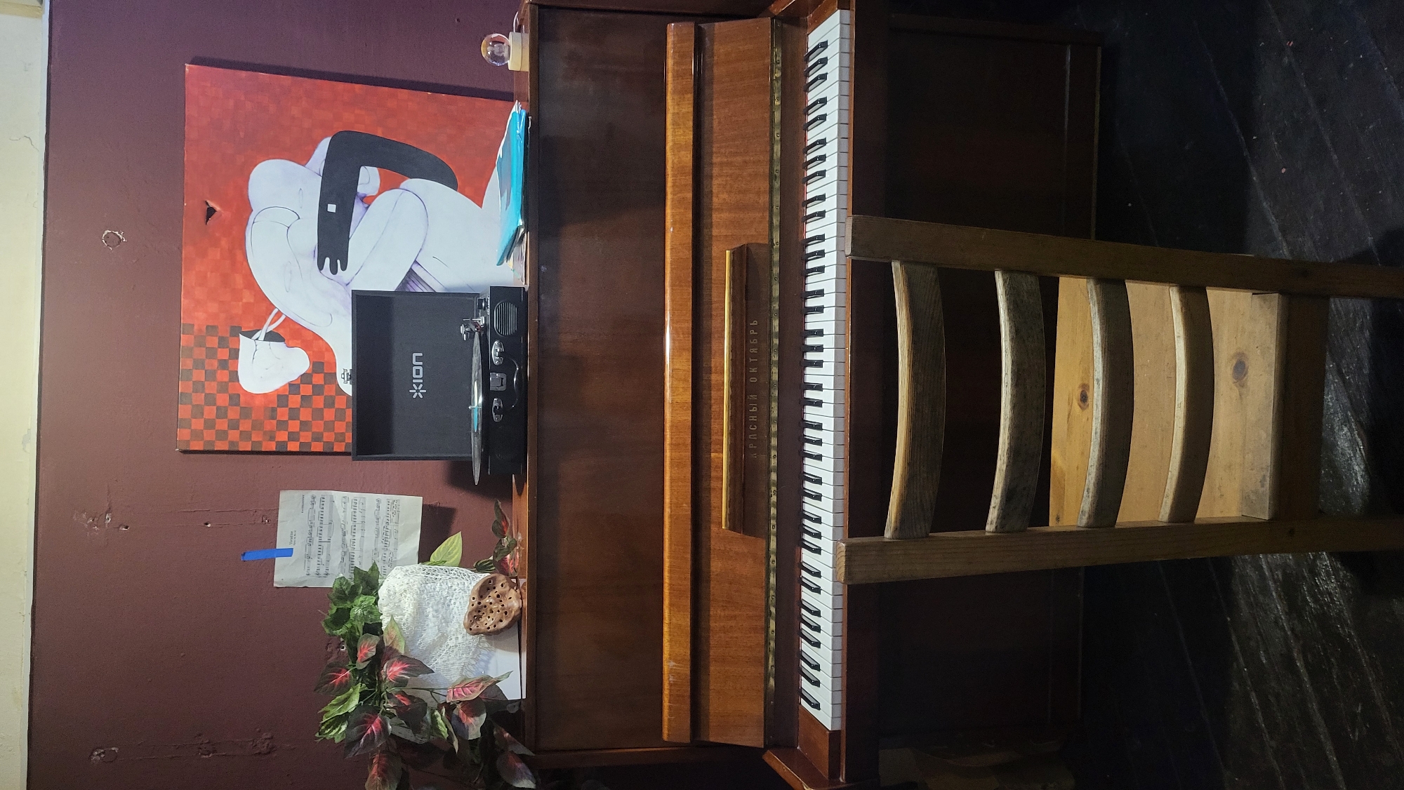 The image shows a wooden upright piano with a chair in front of it. On top of the piano, to the left, is a potted plant and a piece of sheet music attached to the wall with blue tape. To the right of the sheet music is a black record player, and further to the right, a large painting featuring two figures. A small globe light sits on the piano to the far right. The wall behind the piano is a deep red-brown. The piano has a wooden finish and black and white keys. The chair is made of light-colored wood with a slatted back. The floor appears to be dark wood planks. The scene is indoors, likely in a residential setting or a small studio. The location is not specified in the image, and no information about Saint Petersburg, Russia, is present.