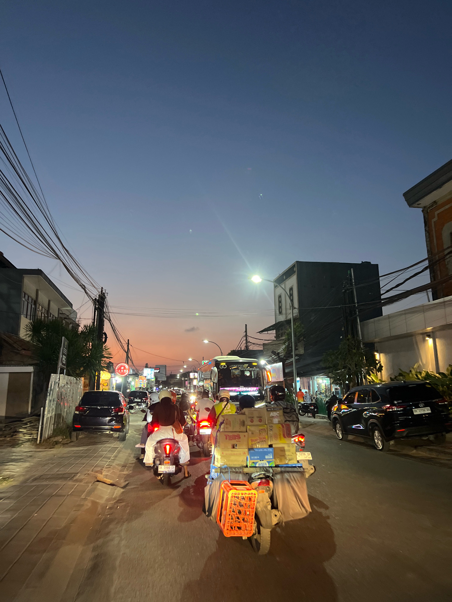 This is a vertical, eye-level, full shot of a busy street scene in Kuta, Indonesia, at dusk. The sky is a gradient of deep blue to soft orange, indicating the transition from day to night. Overhead, a complex network of electrical wires crisscrosses the frame, supported by utility poles.
The street is filled with traffic. In the foreground, a motorcycle with a large cargo carrier is prominent. The carrier is stacked high with boxes of various brands, including "TEBS," "AQUA," and "ABC." An orange plastic basket is attached to the rear of the carrier. The rider and passenger are wearing casual clothing, with the passenger wearing a headscarf.
Several other motorcycles and a black SUV are visible on the road. Some motorcycles have passengers, and the headlights and taillights of the vehicles illuminate the street. The buildings lining the street are a mix of modern structures with multiple floors and some older, more traditional-looking buildings. Many of these buildings have signs and lights, suggesting they are shops or businesses.
The street itself appears to be paved with interlocking bricks in some areas and asphalt in others. The overall atmosphere is one of a bustling urban environment in the evening.