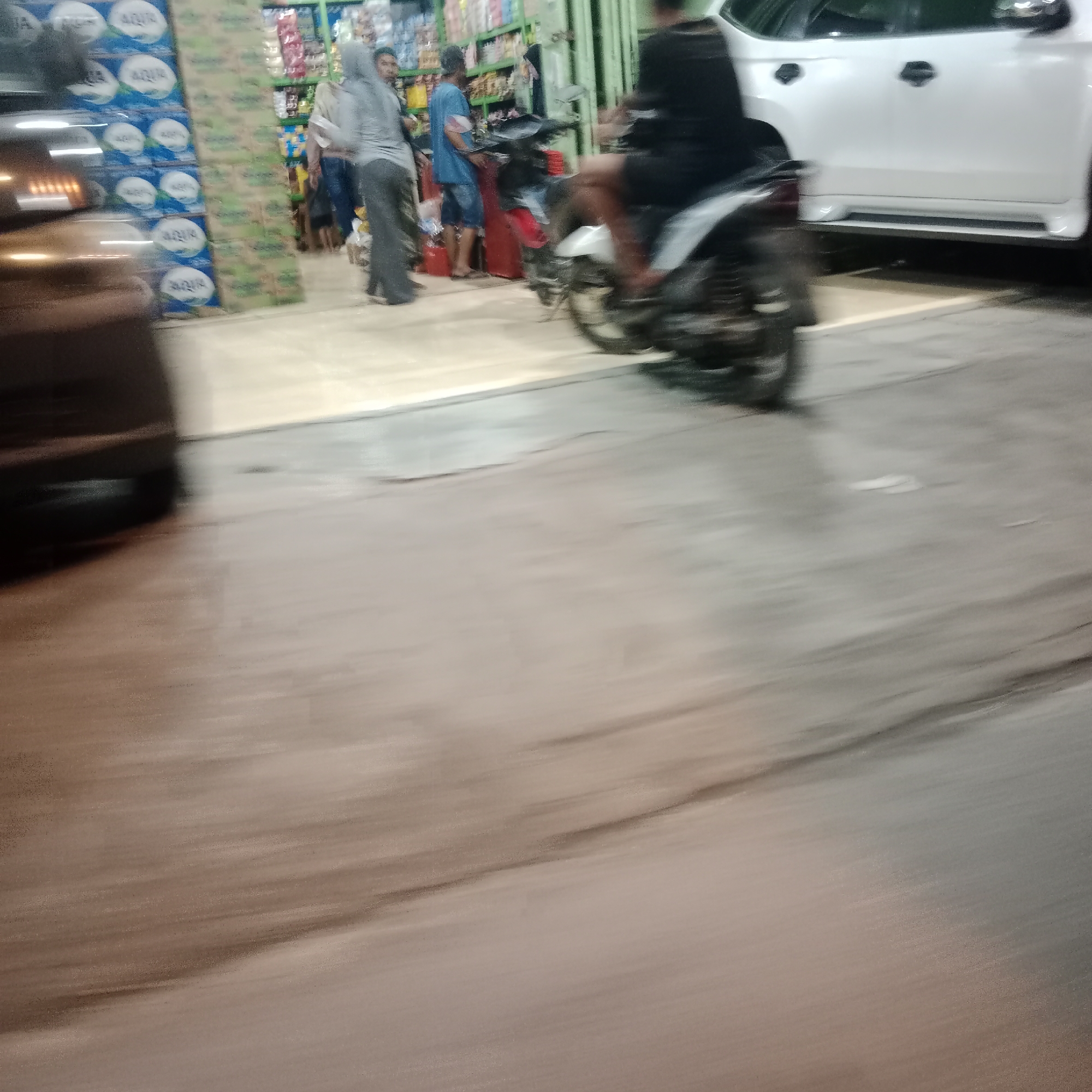 In Gubug, Indonesia, a busy street scene unfolds with motion blur. A white SUV is parked on the right, with a person on a motorcycle partially visible to its left, also in motion. In the background, a store displays shelves stocked with various items, and several individuals, including a person wearing a gray hijab and another in a blue shirt, are visible. The street itself appears to be paved.