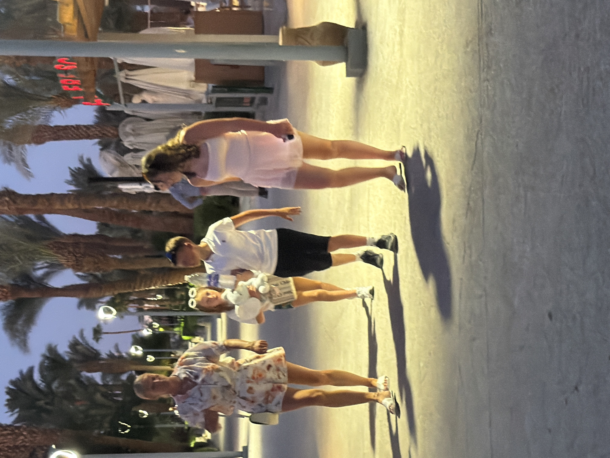In Sharm El Sheikh, Egypt, at dusk, four individuals are walking along a paved promenade lined with palm trees. The woman on the far left wears a floral print romper and white sandals, carrying a white handbag. Next to her, a young girl holds a plush elephant toy and wears a white top and patterned shorts. A young boy walks between them, dressed in a white polo shirt and black shorts, holding a clear plastic bottle. On the right, a young woman in a pink dress walks ahead, looking to her right and holding a mobile phone. In the background, there are covered lounge chairs and a sign with red neon lettering. The lighting suggests it is late evening, with artificial lights illuminating the area.