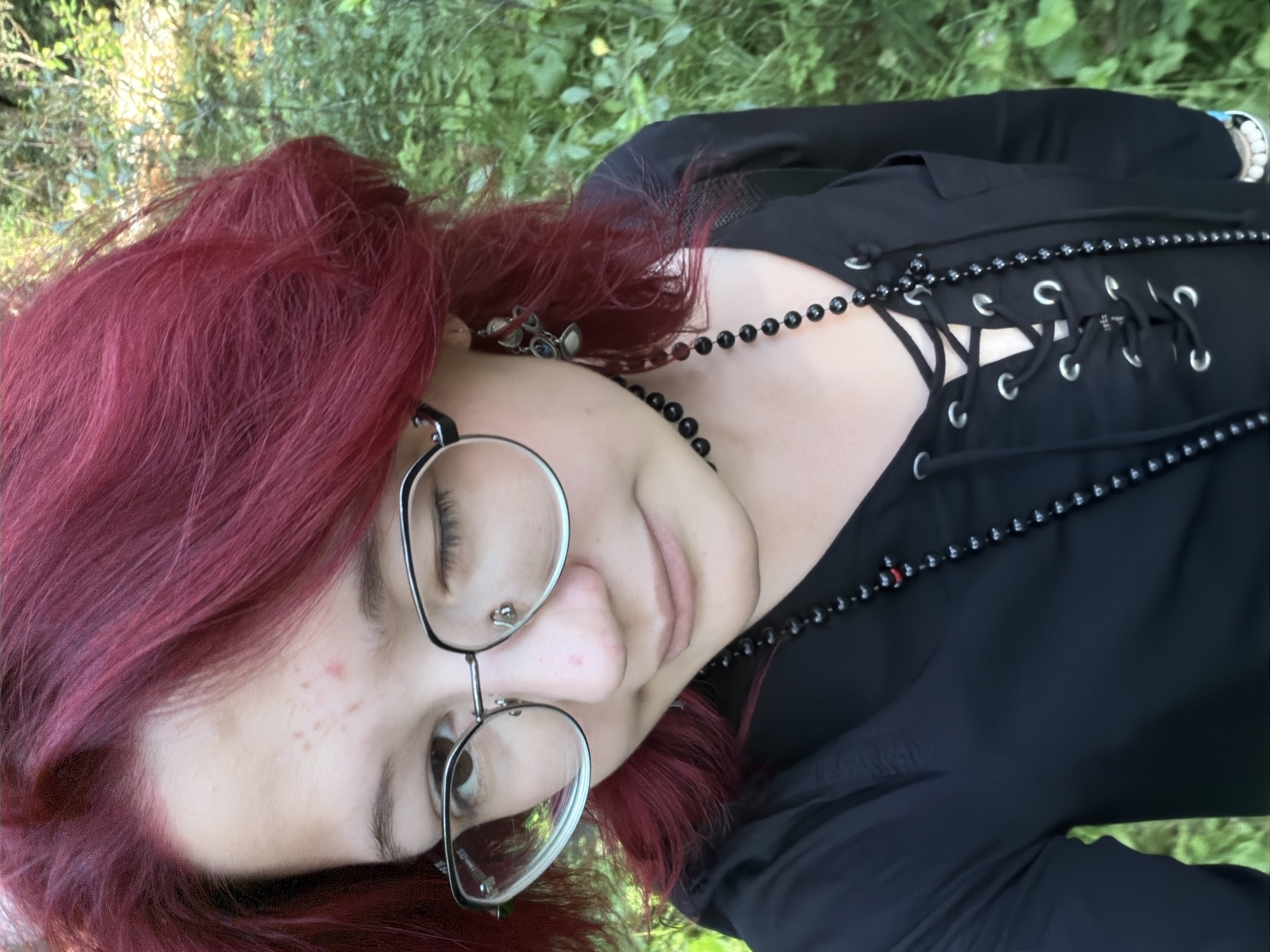 A young person with vibrant red hair and glasses is looking to the side with one eye closed. They are wearing a black top with a lace-up front and multiple necklaces. The background is blurred green foliage, suggesting an outdoor setting. There is no discernible information to place this image in Chelyabinsk, Russia.