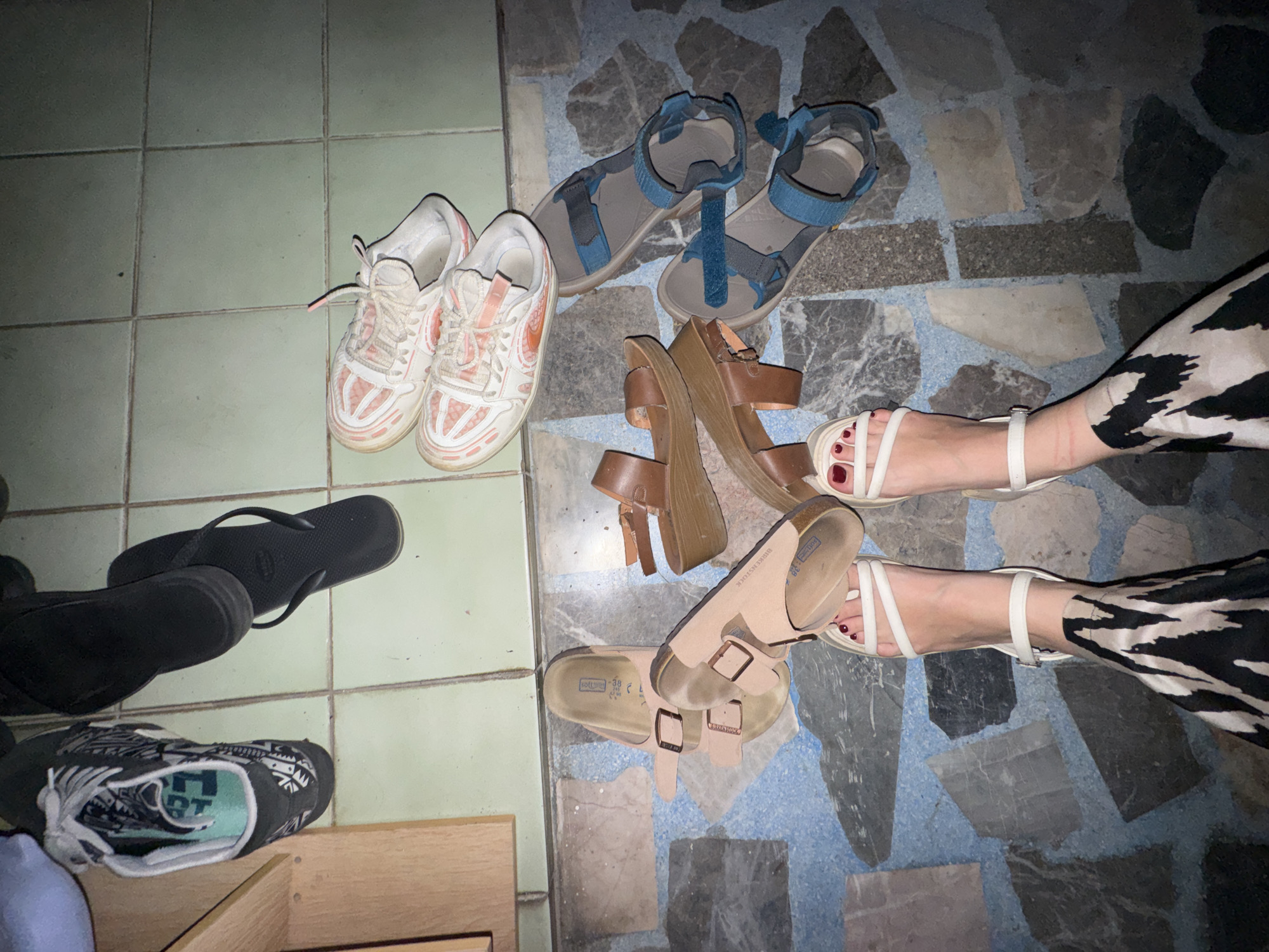 A top-down view shows an assortment of footwear on a tiled floor. Two pairs of sneakers, one black with white and teal accents and another white with pink accents, are positioned on the upper right section of the frame. Next to them, a pair of black flip-flops are visible. In the lower half of the image, a pair of beige Birkenstock sandals lie on their sides, and a pair of brown wedge sandals are stacked. Further down, a pair of blue and grey Teva-style sandals are placed next to each other. At the very bottom, the feet of an individual wearing white strappy sandals with red nail polish are visible, along with a portion of a black and white animal print garment. The floor is a mosaic of light blue, grey, and white tiles. The lighting is dim, casting shadows and obscuring some details. The image appears to have been taken indoors, possibly in a residential setting. There is no indication of the location being in Chiang Mai City Municipality, Thailand.