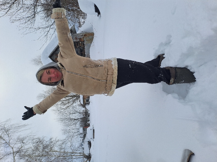 An outdoor, full-body shot depicts an adult female standing in deep snow in Полянки, Russia, under an overcast sky. The subject is positioned with arms raised and extended outwards, palms open, and displays a slight smile. She wears a beige shearling-lined jacket with a zippered front and buckle details, black pants, black gloves, and tall dark boots. A grey hood covers part of her head. Her right leg is deeply embedded in the snow, creating a clear imprint, while her left leg is partially lifted or less visible. The foreground and surrounding ground are covered in undisturbed white snow. In the background, numerous bare deciduous trees are visible, along with several snow-covered rustic wooden structures, including a prominent weathered building with a steeply pitched roof located to the right.