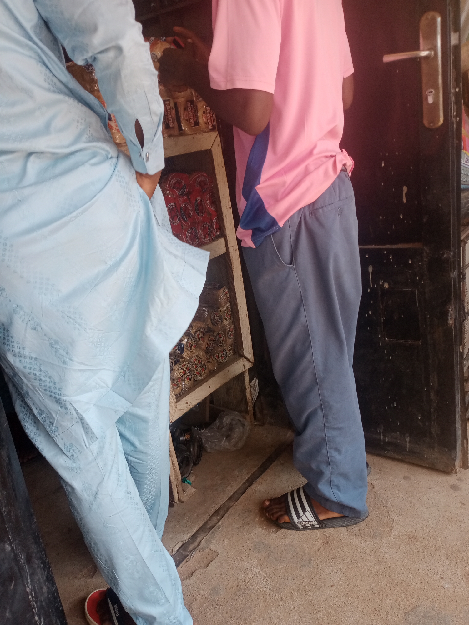 In Jalingo, Nigeria, two individuals are standing near a market stall. The person on the left wears a light blue, patterned traditional garment with long sleeves and visible buttons at the cuff. They are partially obscured by the garment's fabric. The person on the right wears a bright pink t-shirt and dark blue trousers. Their t-shirt has a blue and black stripe detail on the side. They are wearing black and white striped slide sandals. The stall behind them contains shelves stocked with various packaged food items, including what appear to be biscuits and other snacks. A metal door with a handle and lock is visible to the right of the frame, and the ground is a rough concrete surface. The scene suggests a commercial setting within the city.