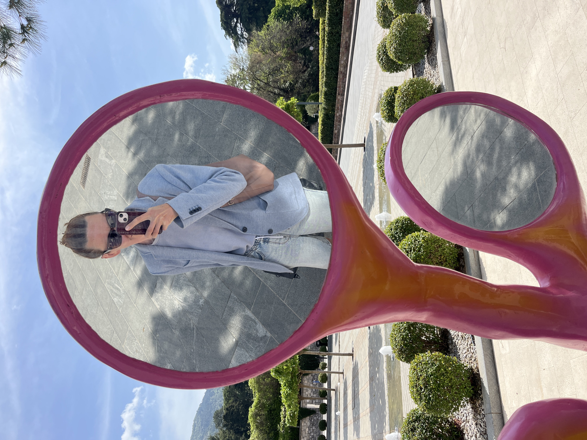 The image depicts a person taking a selfie in a large, pink, decorative sculpture resembling a pair of scissors. The scissors appear to function as mirrors, reflecting the person and the surrounding environment. The person is wearing sunglasses, a light blue blazer, and light-colored jeans, holding a smartphone.
The location appears to be a landscaped area in Tivat, Montenegro, characterized by meticulously trimmed shrubs, trees, and a patterned stone pavement. Small fountains are visible in the background, suggesting a well-maintained public space. The overall environment suggests a sunny day.