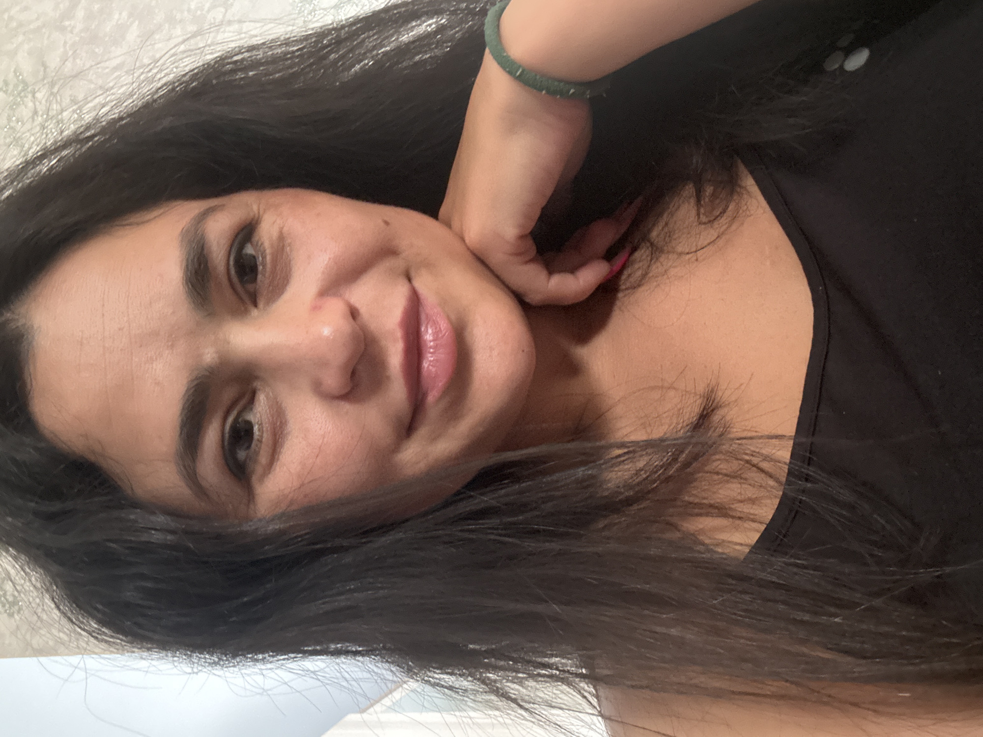 A woman with long, dark hair and a light complexion is looking at the camera and smiling slightly. She is wearing a black top and a green beaded bracelet on her right wrist. Her right hand is touching the side of her neck/jaw. The background is blurred, but appears to be a light-colored wall. There is no evidence to suggest the location is in Кемуль, Russia.