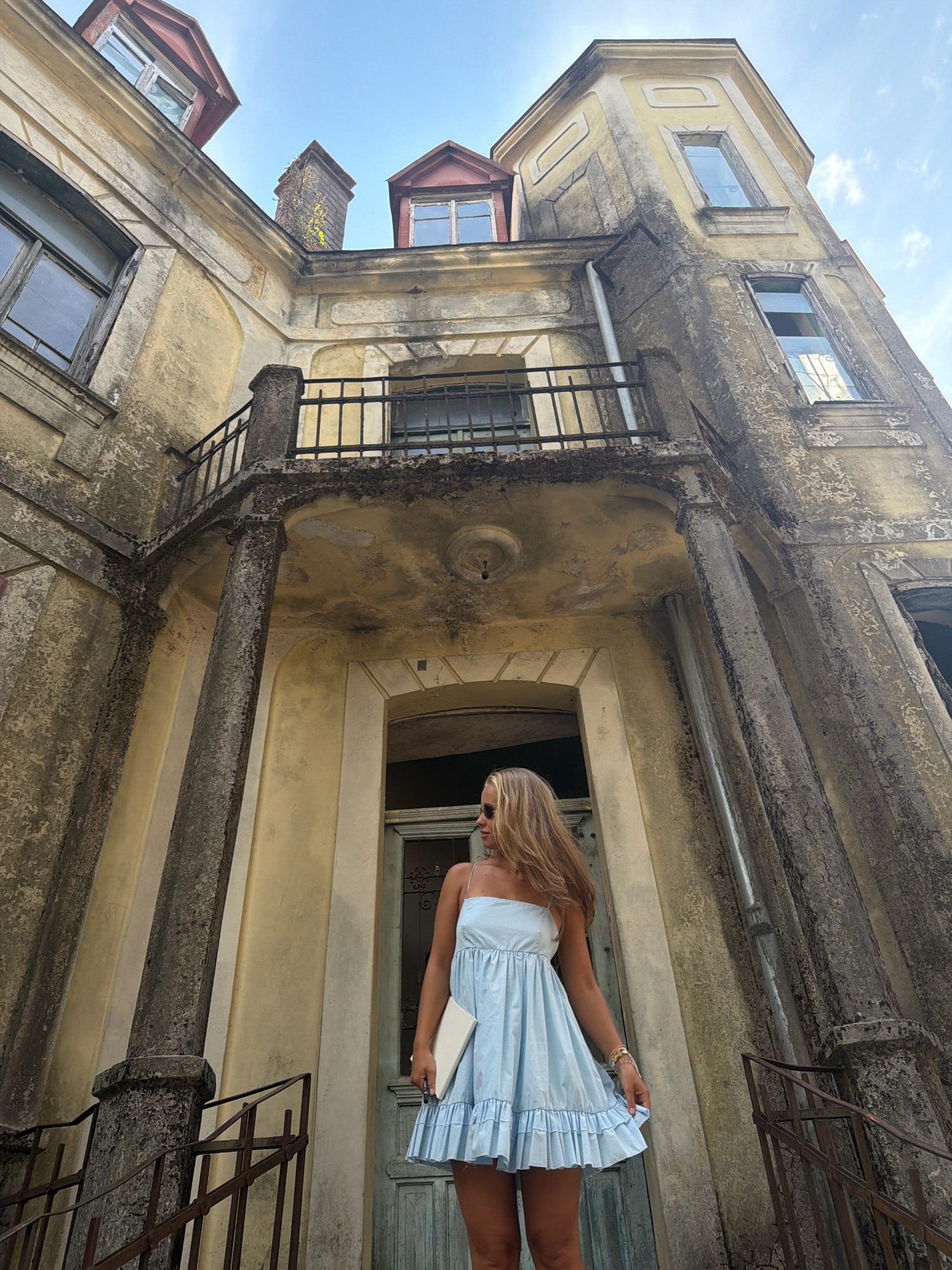 A low-angle shot captures a young woman standing in front of a dilapidated yellow building with weathered stone columns and balconies, likely in Cetinje, Montenegro. The woman, with blonde hair and sunglasses, wears a light blue ruffled dress and holds a white clutch. She is looking to her left. The architecture suggests an old, possibly abandoned, estate, with peeling paint and cracked plaster on the building's facade. The sky is visible above, with hints of clouds. Stairs with rusty railings lead up to the building.