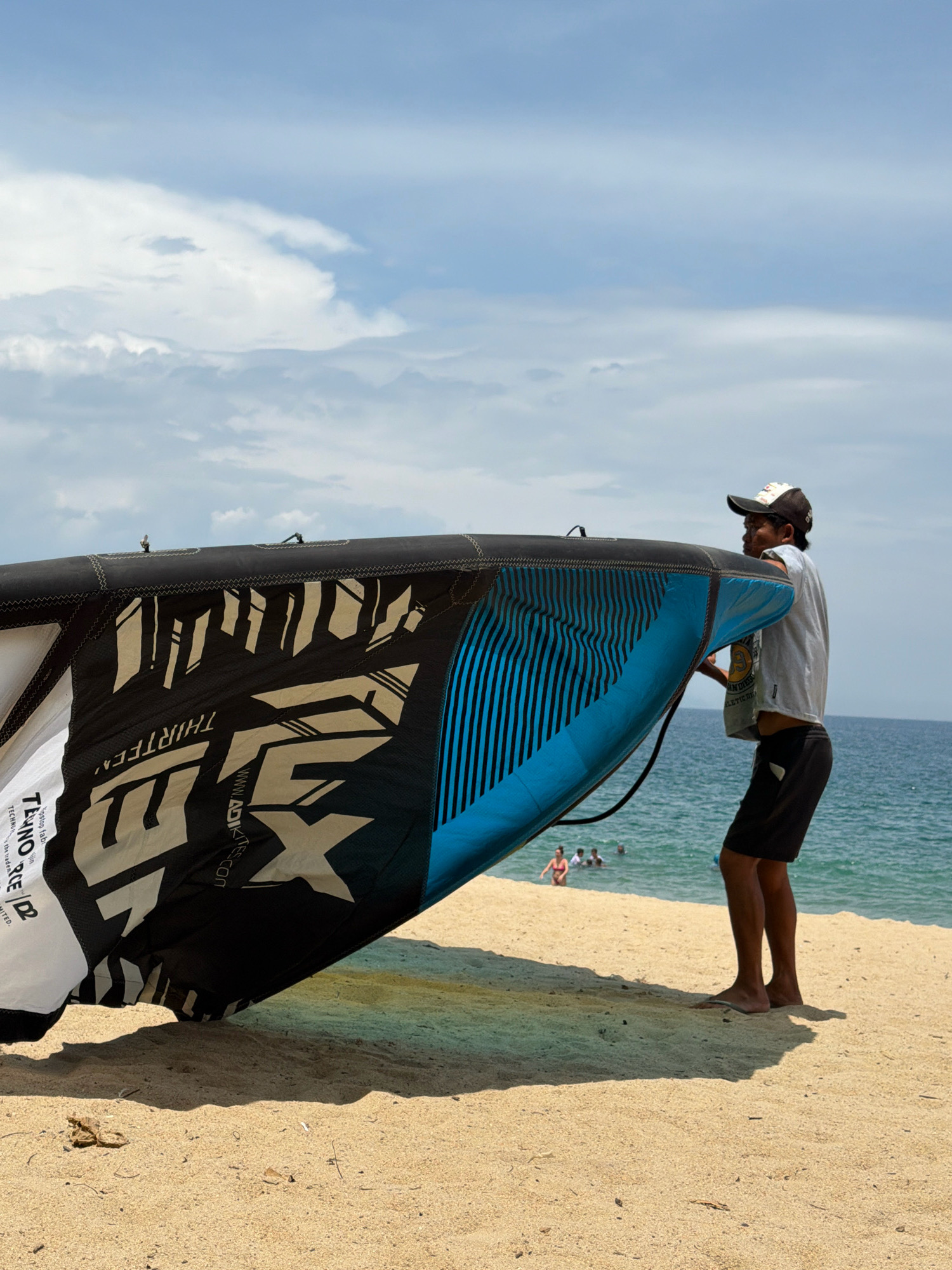 An overcast day at a beach in Nha Trang, Vietnam. A man wearing a cap, grey t-shirt, and black shorts is holding a large, black and blue kite. The kite has white and blue graphics on it, including text that reads "THIRTEEN" and "ADIKITES.COM". The kite is partially unrolled, with the blue section facing the viewer. The man is standing on the sandy beach, with the ocean in the background. Several people are in the water, swimming. The sky is mostly cloudy with patches of blue.