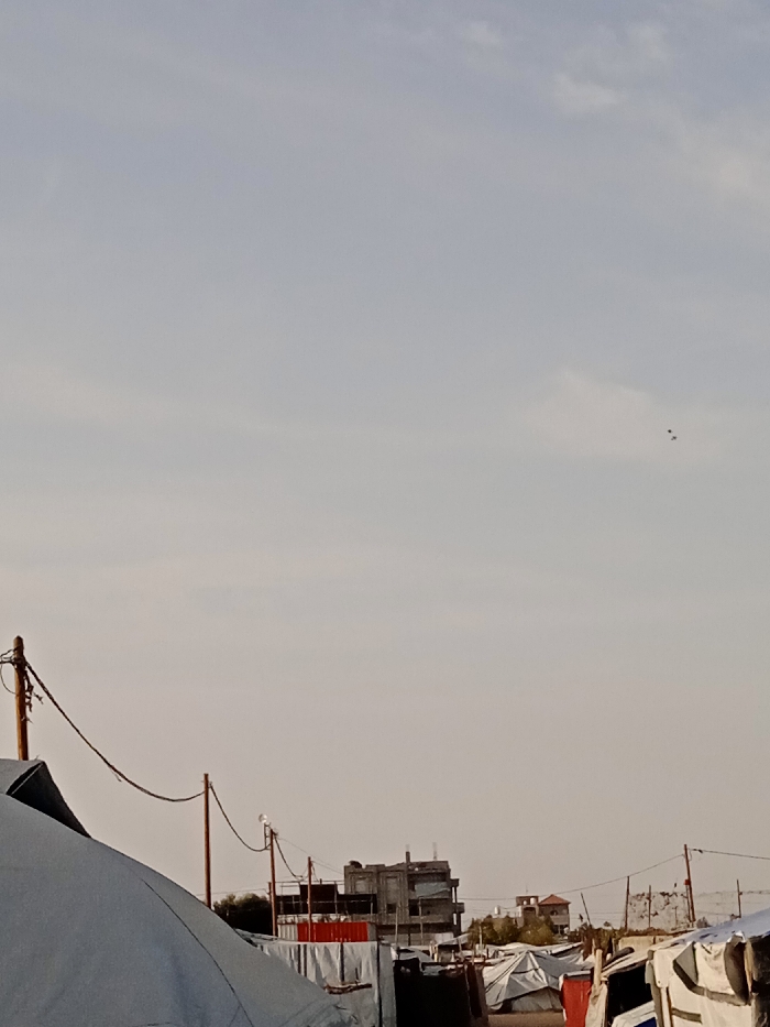 A wide shot shows a landscape filled with tents and makeshift shelters, possibly a refugee camp or impoverished area. Utility poles with wires cross the foreground. A partially constructed or damaged building is visible in the middle distance. The sky is pale and hazy, with a few small birds or objects in the far distance. The overall scene suggests a temporary and possibly challenging living situation.
