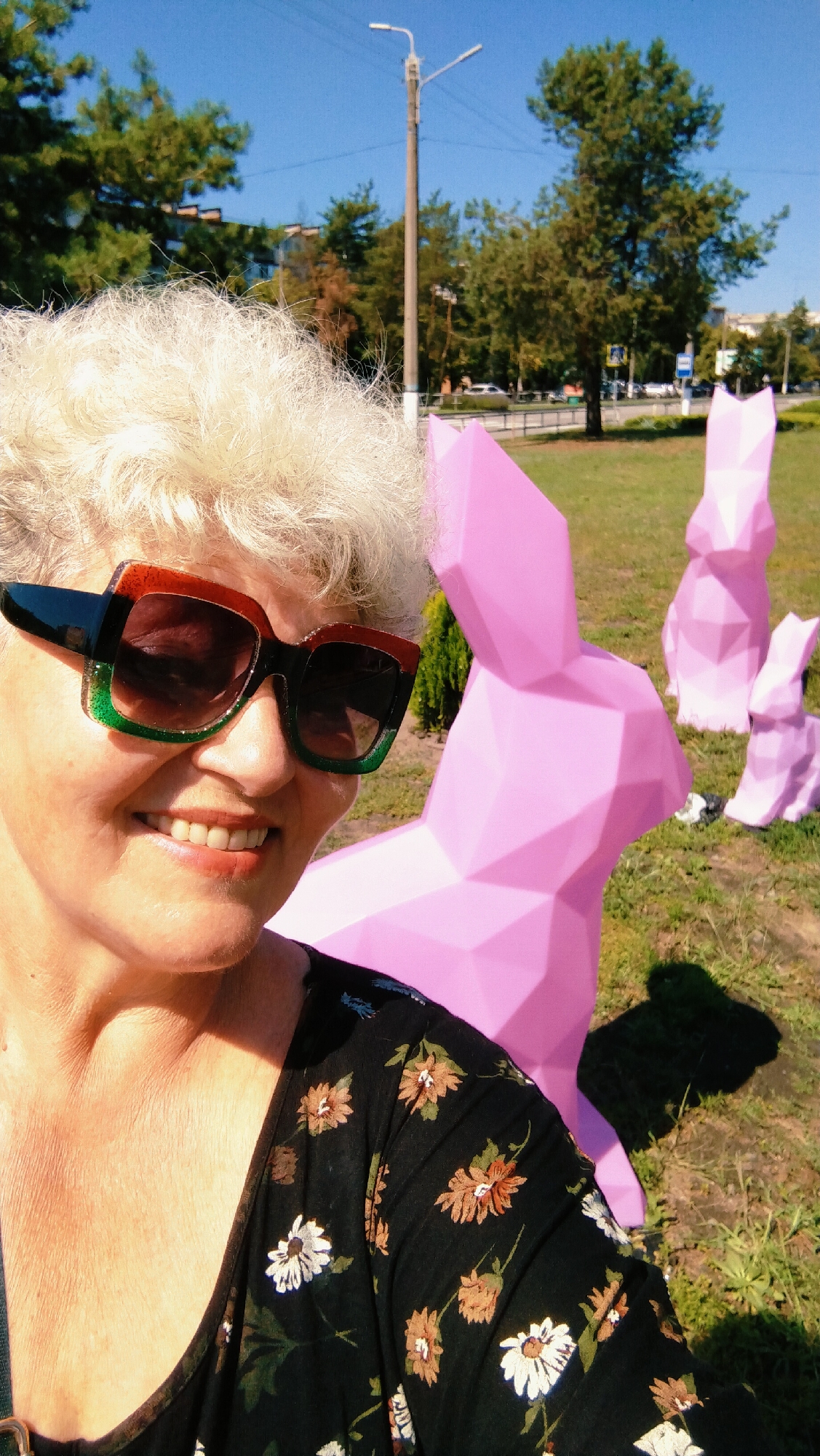 A woman with short, curly, light-colored hair and sunglasses with thick frames, which are orange and green, takes a selfie outdoors. She is wearing a black dress with a floral pattern featuring white and orange flowers. The background includes green grass, trees, a street with cars, buildings, a lamppost, and several large, pink, polygonal rabbit sculptures. The image appears to be taken in a park or public space in Horishni Plavni, Ukraine, given the context provided. The sunlight casts shadows on the grass, indicating it is daytime.