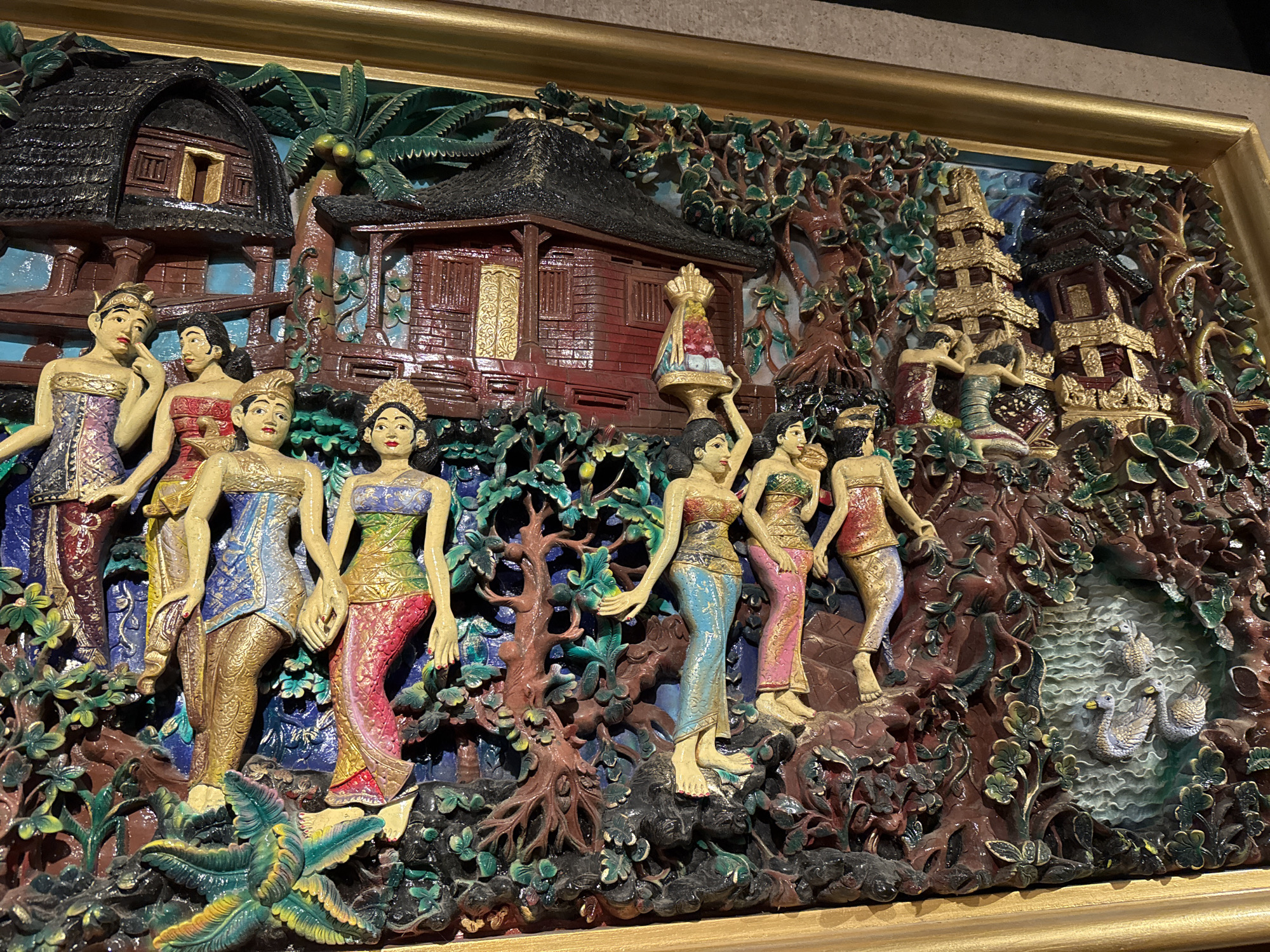 The image displays a meticulously carved wooden bas-relief, framed by a simple gold border. The artwork depicts a vibrant scene with multiple figures and structures, rendered in rich, painted colors. In the background, traditional Indonesian houses with thatched roofs and ornate details are nestled amongst lush greenery and palm trees. To the right, a multi-tiered pagoda-like structure stands tall, surrounded by dense foliage.
The foreground is populated by several carved figures of women, adorned in brightly colored, traditional clothing, featuring intricate patterns and gold accents. They are depicted in various poses, some standing and some with bent knees, engaged in activities that suggest a communal gathering or daily life. One woman in the center is carrying a tiered, decorated object on her head.
The environment is detailed with various plants, trees, and a water feature with carved ducks. The overall impression is one of a lively and culturally rich setting. The style of architecture and the attire of the figures suggest a Balinese influence, potentially representing a scene from a village in Kuta, Indonesia, a region known for its vibrant arts and crafts.