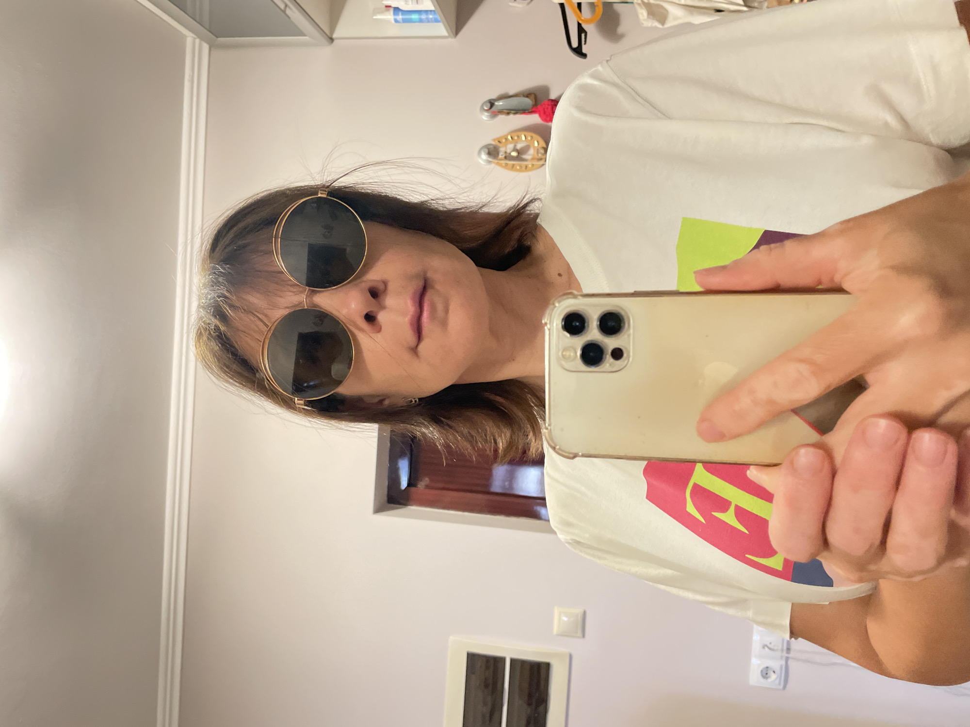 An adult female, estimated to be middle-aged, is depicted indoors taking a self-portrait with a smartphone. She has shoulder-length brown hair with visible grey roots and wears large round sunglasses with gold frames, obscuring her eyes. Her lips are slightly parted. She is dressed in a white short-sleeved t-shirt featuring a colorful graphic design with prominent pink, lime green, and purple shapes, including a lime green capital "F." She holds a light-colored smartphone, consistent with an iPhone model featuring a triple camera array, protected by a clear case.
The background shows an interior room, presumed to be in a residence in the city of Sochi, Russia. The walls are a light, neutral color, and the white ceiling features decorative crown molding. A ceiling light fixture is reflected in the sunglasses. On the left side of the frame, a white rectangular wall decoration is mounted above a white electrical switch. Further left, a white double electrical outlet is visible. A dark wooden doorframe or door is partially visible through an opening. On the right, white shelving units hold various bottles. Below the shelves, two decorative wall hooks are present; one supports a yellow, studded horseshoe-shaped ornament, and the other holds a grey and pink object. A dark clothes hanger with a yellow hook is visible further right.