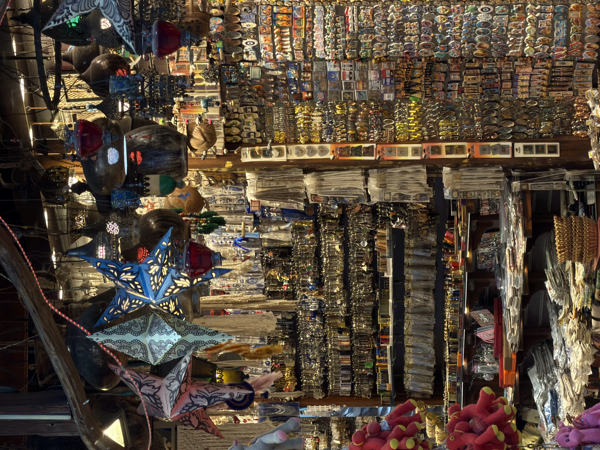 This media file captures an indoor marketplace scene, likely a souvenir shop in Sharm El Sheikh, Egypt, judging by the abundance of trinkets and decorative items. The image is an overhead shot, with the perspective tilted, making it feel as though we are looking down into a bustling, vertically oriented space.
The primary subjects are a vast array of objects. Hanging from the ceiling and shelves are numerous star-shaped lanterns, some illuminated from within. These lanterns feature intricate patterns in various colors, including blue, white, and pink. Further into the scene, shelves are densely packed with merchandise. We can see rows upon rows of small, colorful items that appear to be magnets or small decorative pieces, many with images and text. There are also stacks of what look like postcards or small prints, some in plastic sleeves. In the foreground, piles of red, plush toys with yellow accents are visible, as are some grey and pink plush toys in the lower left. The overall impression is one of a shop overflowing with goods, designed to attract tourists.
There are no people visible in the image, nor is there any indication of a specific time of day or weather conditions, as it is an indoor setting. The sheer volume and variety of souvenirs suggest this is a tourist-oriented location. The lighting is somewhat dim, with spotlights illuminating some of the hanging items, creating a vibrant and somewhat cluttered atmosphere. The dominant emotion conveyed is one of abundance and perhaps a touch of sensory overload, typical of a souvenir market. The location cue "Sharm El Sheikh, Egypt" strongly suggests a context of holiday shopping and mementos from a popular tourist destination.