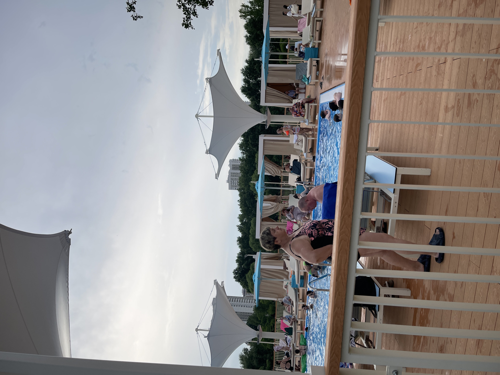 The image depicts a crowded outdoor swimming pool area on a cloudy day. In the foreground, a wooden deck with white railings is visible, with people standing and sitting. A woman in a floral swimsuit stands near the railing, facing left. Next to her, a man with a blue towel on his lap is seated. In the pool, several people are swimming and interacting. Behind the pool, lounge chairs and cabanas are set up, with more people relaxing. Two large white tensile structures, common in resort settings, provide shade. In the background, trees and tall buildings are visible, suggesting an urban environment. The presence of lounge chairs, cabanas, and the swimming pool points to a recreational facility, possibly a water park or a hotel pool area. The Moscow skyline in the background, with its distinctive architecture, helps to locate the scene.