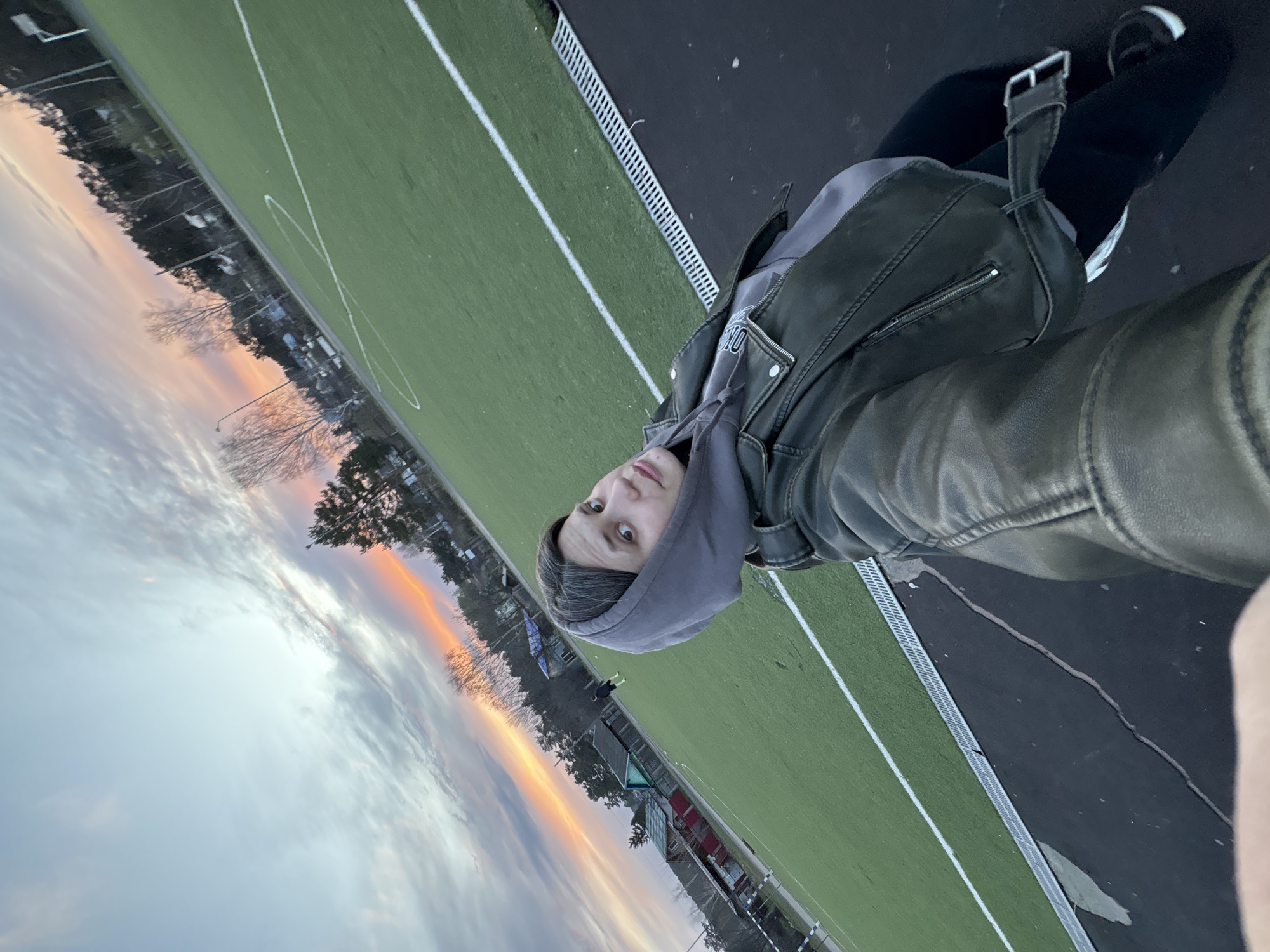 This image, taken in Chaikovsky, Russia, depicts a person taking a selfie at an outdoor sports field during sunset or dusk. The photograph is rotated ninety degrees clockwise.
In the right foreground, the subject looks directly into the camera with a neutral expression. They are wearing a grey hooded sweatshirt under a dark, possibly leather, jacket. Their attire also includes dark leggings or pants and dark athletic shoes with white soles or accents. A dark shoulder bag with a distinct buckle strap is visible hanging from their shoulder.
The setting is a green artificial turf sports field, marked with white lines, reminiscent of a soccer or football pitch. To the right of the field, there is a dark asphalt or track area, which features a long metal grate, likely part of a drainage system.
The background showcases a vibrant sunset sky, with warm orange, pink, and purple clouds merging into lighter grey and blue tones. Silhouettes of bare and evergreen trees are visible against the colorful horizon, alongside distant buildings and a street light, suggesting a semi-urban or residential area bordering the field. The overall ambiance is peaceful. No visible text is present in the image.