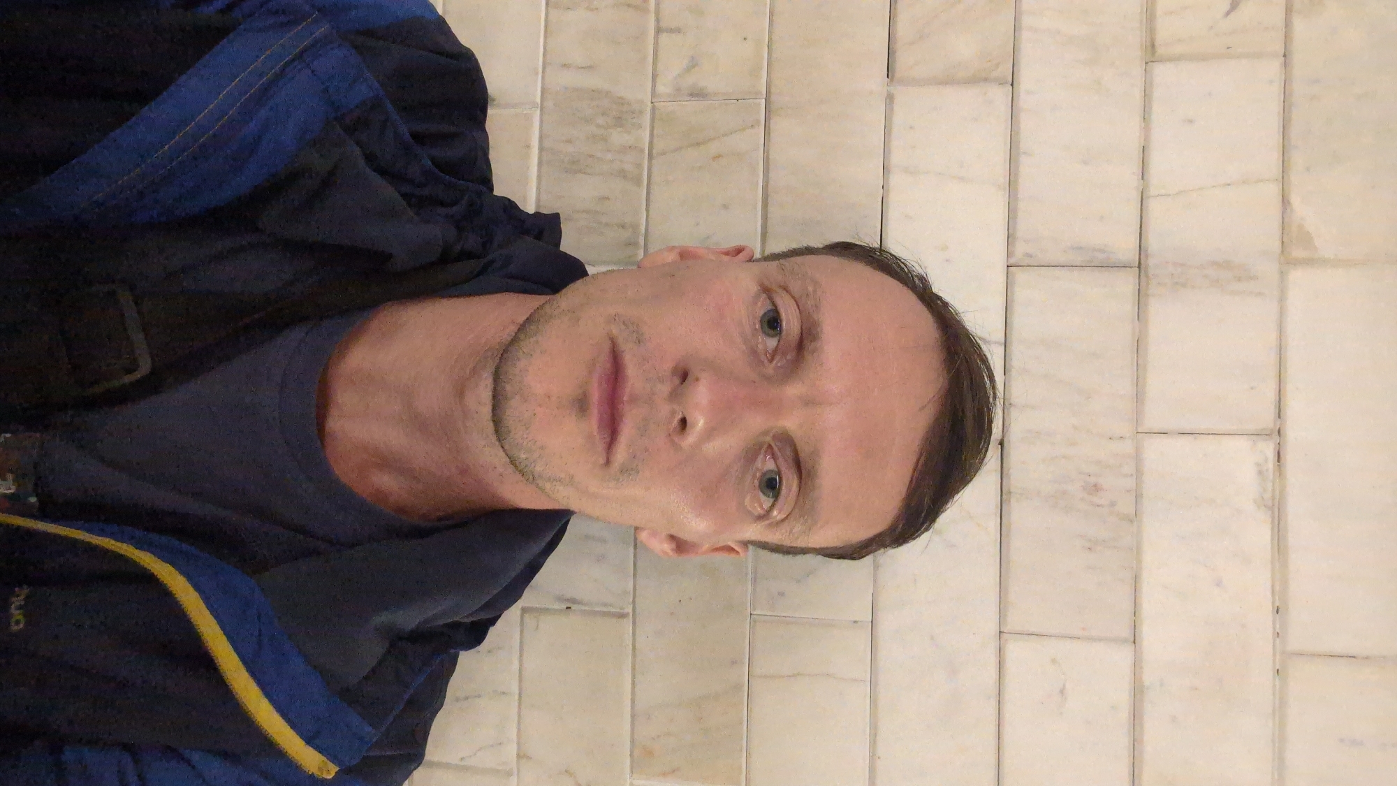 A man is posing for a selfie indoors, angled slightly. He has fair skin, short brown hair, and blue eyes. He is wearing a dark blue T-shirt and a dark blue jacket with yellow trim. The background is a wall tiled with rectangular, cream-colored marble tiles. There is no indication of a location in Da Lat, Vietnam.