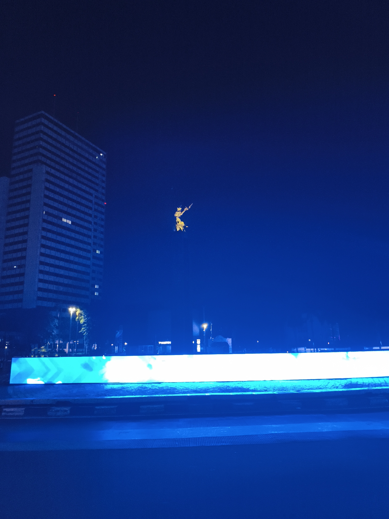 The image captures a nighttime scene with a tall, modern building on the left side of the frame. In the center, there's a statue or sculpture in mid-air, illuminated in yellow. Below, a long LED display stretches horizontally, emitting a bright white light. The foreground is a dark blue surface, possibly a road or walkway. The city is Unknown, Unknown.