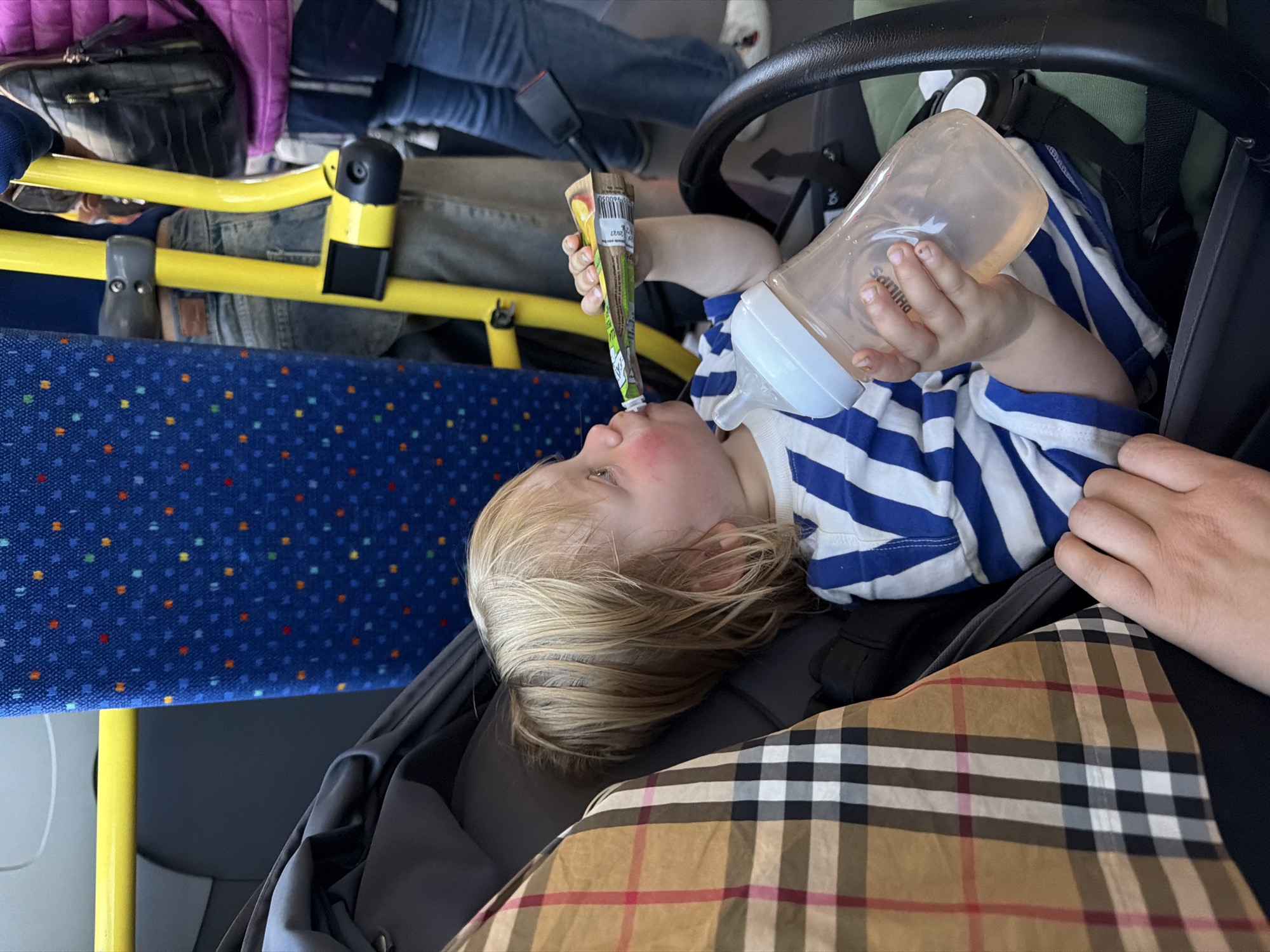 The image shows a child lying in a stroller on what appears to be a public bus, evident from the patterned blue seating and yellow handrails. The child is blond with fair skin, wearing a blue and white striped shirt, and drinking from a squeezable fruit pouch. They also hold a baby bottle. A hand is partially visible on the right edge of the frame. Other people's legs and belongings can be seen in the background. The overall setting suggests a scene of everyday life, and assuming the accuracy of the specified location, the bus would be located in Capiago Intimiano, Italy.