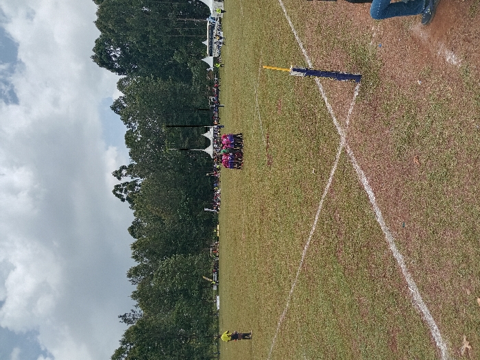 An outdoor sports event is depicted on a partially worn grass field with white painted lines, under a cloudy sky in Meru, Kenya. In the central mid-ground, a huddle of approximately 10-15 individuals in matching purple and pink uniforms is visible, likely athletes. To the far left mid-ground, a lone individual in a yellow shirt and dark trousers stands observing. In the background, numerous spectators are gathered near several white canopy tents and a white bus or large van. A pair of black goalposts, typical for rugby, stands in the distance, centered behind the huddle. The foreground shows an intersection of white lines on the grass, with a blue and yellow marker pole positioned upright near the lower right frame, adjacent to the visible lower legs of an individual seated or standing outside the main playing area. The field is bordered by a dense line of tall green trees.