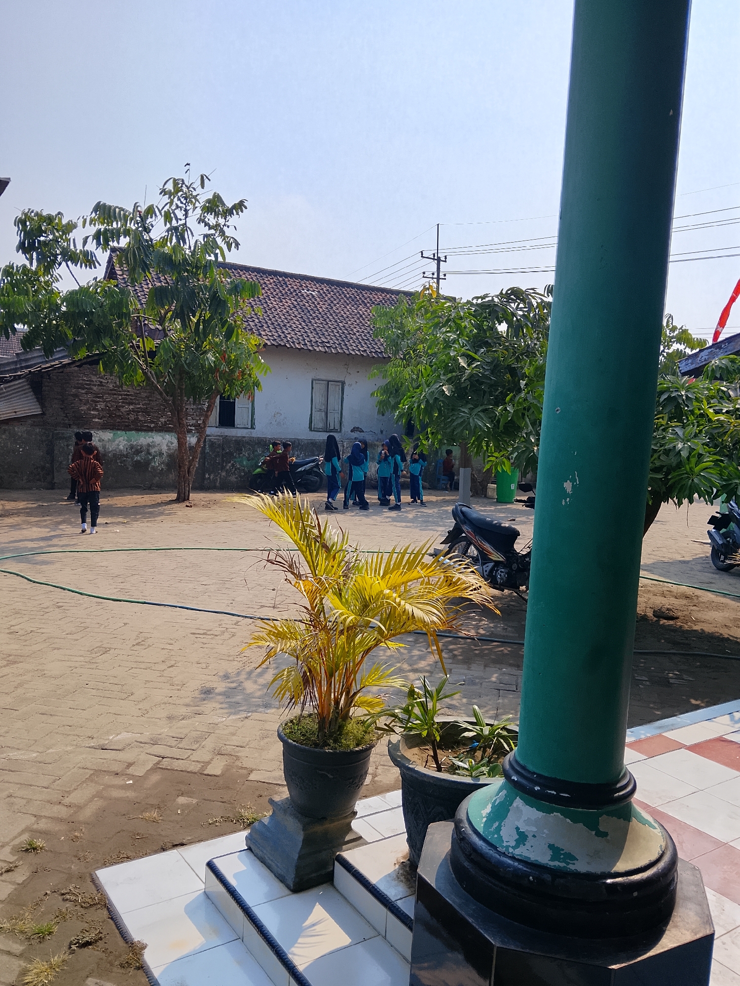 This image depicts an outdoor scene in what appears to be a schoolyard or open public space in Kepuh, Indonesia. The foreground is dominated by a large, green, cylindrical pole, possibly a flagpole, with a chipped paint finish. To the left of the pole, two potted plants are visible. The larger plant has yellow-green, spiky leaves, while a smaller plant with green leaves stands in the adjacent pot. The ground is paved with interlocking tan bricks, with patches of dry dirt and sparse weeds.
In the mid-ground, a group of young individuals, likely students, are gathered. Some are wearing uniforms: two girls in blue and teal hijabs and dresses, and one boy in a patterned brown and white shirt and dark pants. To the left of this group, another boy in a similar patterned brown and white shirt and dark pants stands alone. Behind them, a single-story building with a white facade, a reddish-brown tiled roof, and visible windows with shutters is present. Trees with green foliage are scattered throughout the mid-ground and background, suggesting a tropical or subtropical climate. Two motorcycles are parked near the building, one black and the other partially visible. The sky above is clear and bright blue, indicating a sunny day. The overall impression is of a typical Indonesian school environment during daylight hours.