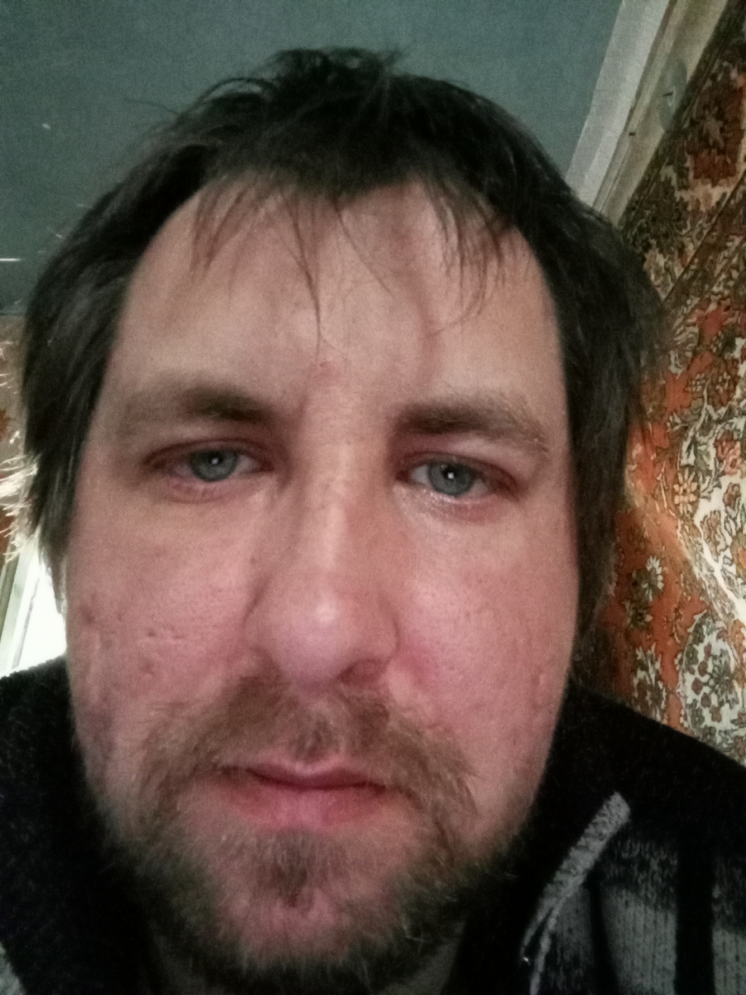 A close-up shot of a middle-aged man's face. He has fair skin, blue eyes, and dark hair with some graying. He has a beard and mustache. His face shows signs of acne scarring. He is wearing a dark-colored, possibly wool, sweater. The background includes a pale green wall and a patterned carpet or tapestry, suggesting an interior setting, likely a home. The lack of external cues prevents confirming that the photograph was taken in Parabel, Russia. There is no interaction or action evident in the photo, it is a static portrait.