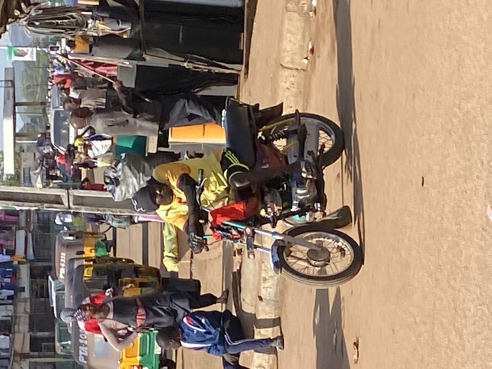 This image captures a vibrant and bustling street scene in Bukuru, Nigeria, under clear, sunny daylight. The ground is a mix of dirt and pavement, separated by a curb.
In the foreground, a man wearing a yellow top and dark shorts is seated on a black and silver motorcycle, facing away from the camera. His feet are on the ground, and he appears to be waiting or resting. To the right, a young boy in a blue and white striped jacket and dark pants is kneeling on the ground.
The midground and background are densely populated with numerous people and vehicles, indicating a busy commercial or market area. Several three-wheeled auto-rickshaws, commonly known as keke napep in Nigeria, are visible; some are yellow, green, or brown. Visible text on these vehicles includes "PM-6258" and "PTA-10...", likely license plates. A white van or bus is further in the distance.
People of various ages are walking, standing, and interacting. Many appear to be vendors with goods piled up or displayed near stalls, although the specific items are indistinct due to distance and resolution. The overall atmosphere is one of active daily life in an urban setting. Shadows are distinct and relatively short, suggesting a mid-day sun. The scene is full of movement and everyday activities typical of a West African street market.