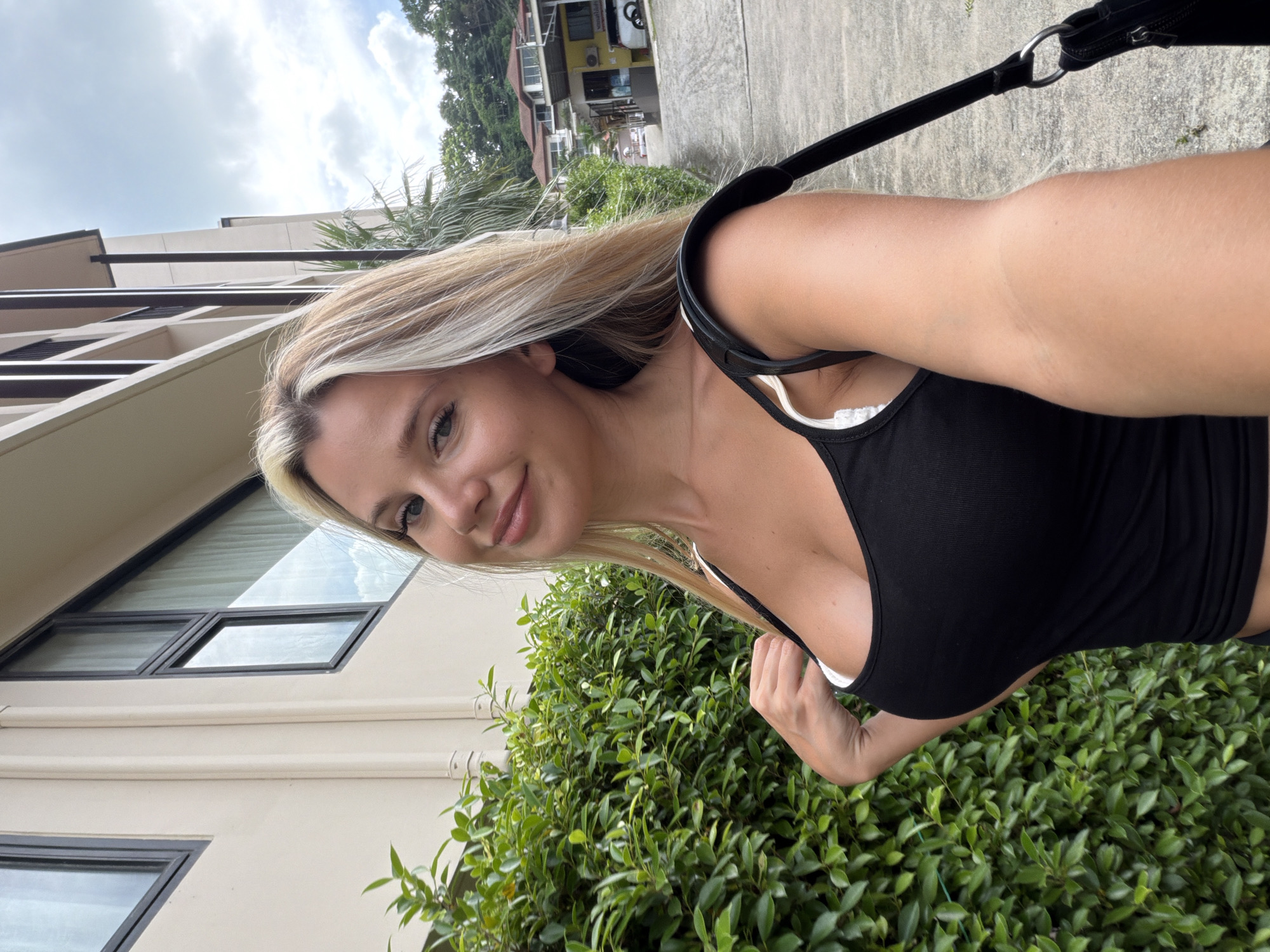 An outdoor, eye-level, selfie-style medium shot features a young, fair-skinned woman with blonde hair, posing in front of a building. She is wearing a black tank top with a white bra visible underneath. Her right hand is resting on her chest. To her left is a dense green hedge, and behind her is a paved path leading to buildings and trees. The setting appears to be a tropical or sub-tropical location, consistent with the city of Wichit, Thailand, known for its resorts and lush vegetation.