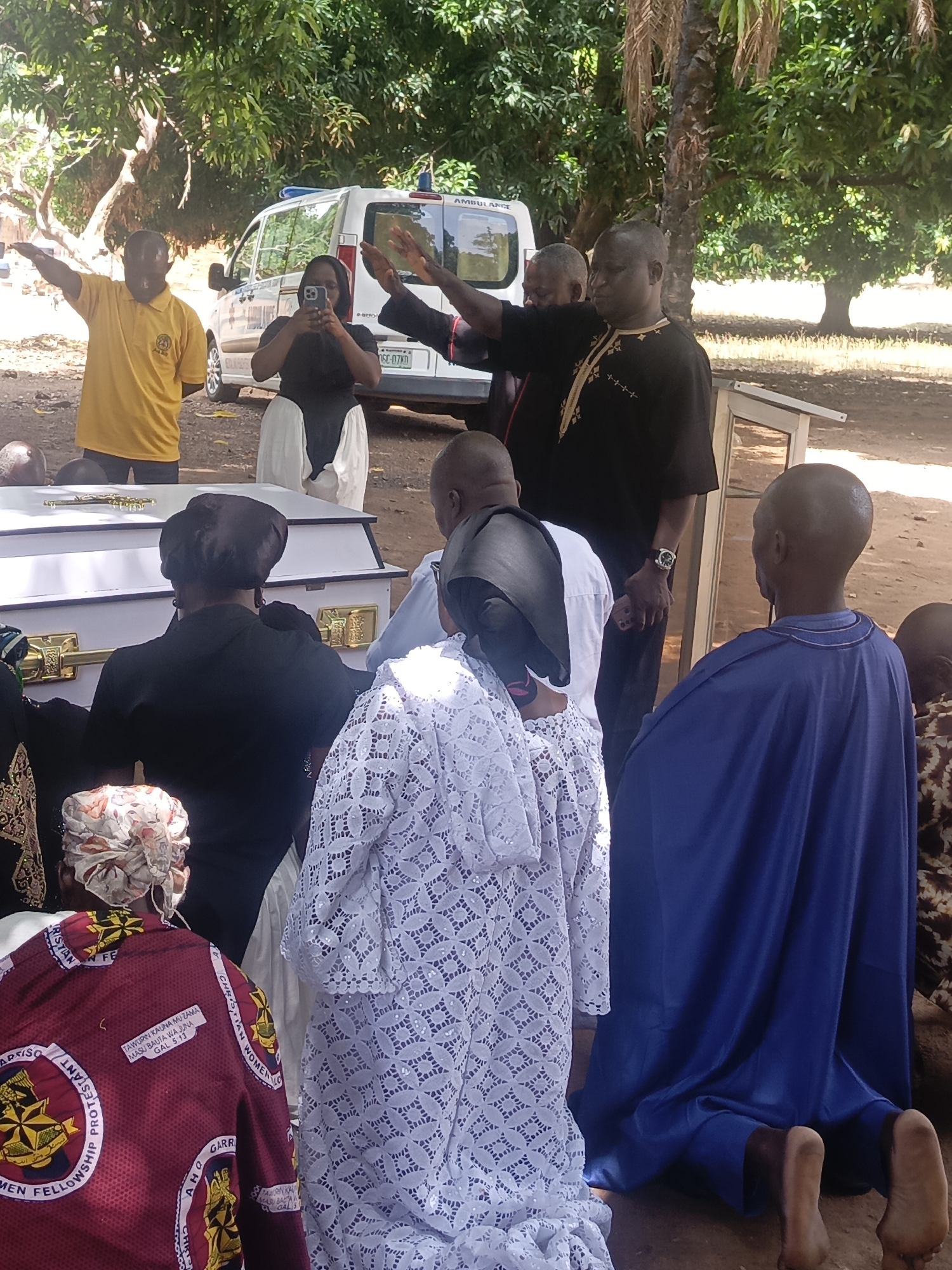 A funeral service is taking place in what appears to be Ninzo South, Nigeria. The scene includes a white coffin with gold accents, around which mourners are gathered. Some mourners are kneeling, while others are standing. Individuals are wearing a variety of clothing including white lace, black attire with gold embroidery, yellow shirts, and blue fabric. A person is taking a photo with a phone. An ambulance is parked in the background. The scene is under trees and in an outdoor environment. A plate on the car says "06C-07KD".